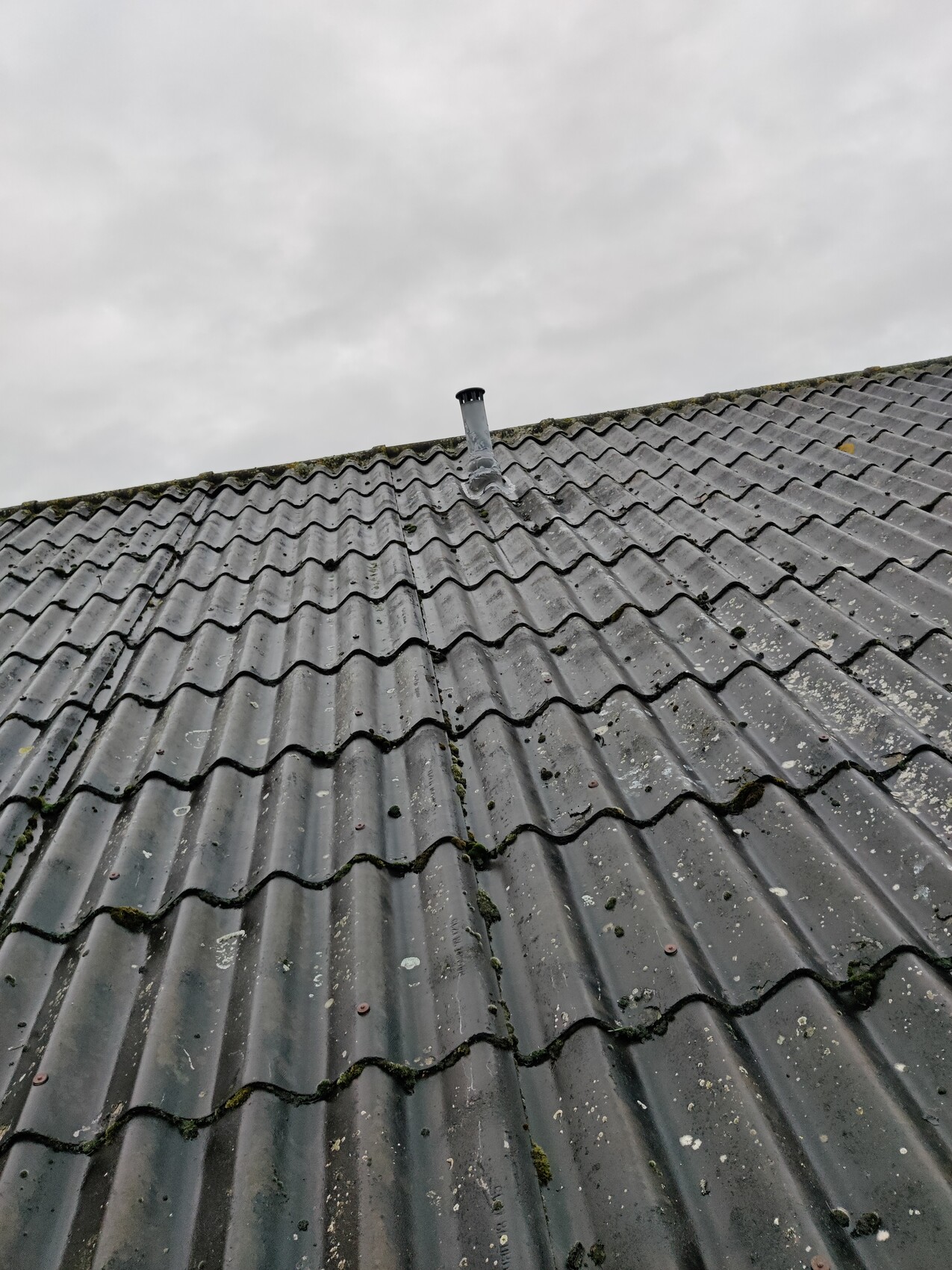 Graues Dach mit gewellten Ziegeln, Moos an Fugen, Kaminrohr in der Mitte, wolkiger Himmel.