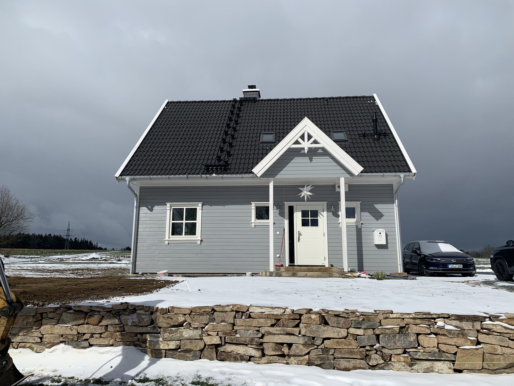 Kleines graues Einfamilienhaus mit weißem Eingang, schwarzem Dach, Schnee und Steinmauer davor.