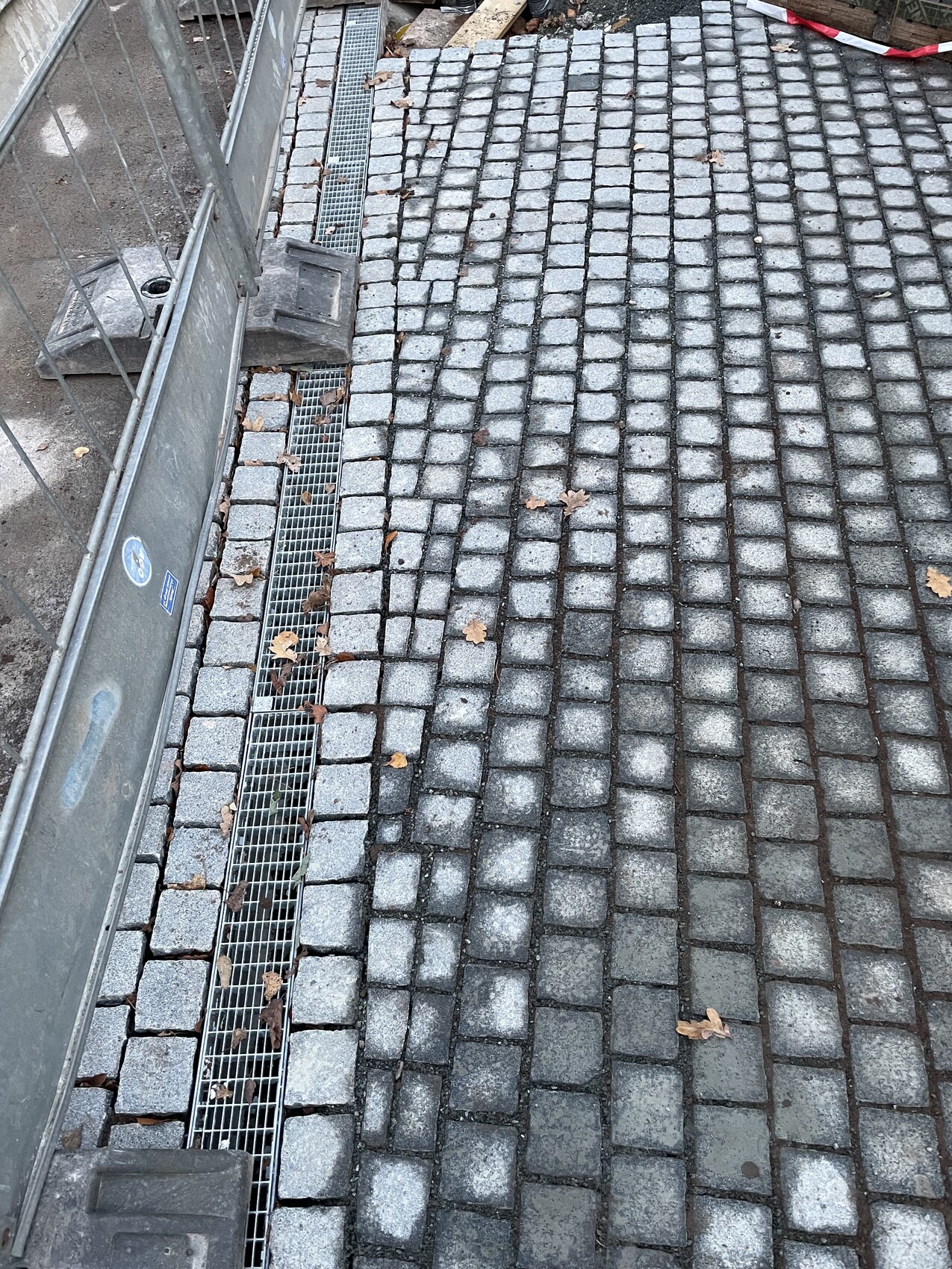Grauer Gehweg aus Kopfsteinpflaster mit Metallrinne am Rand, links Bauzaun, Herbstlaub.