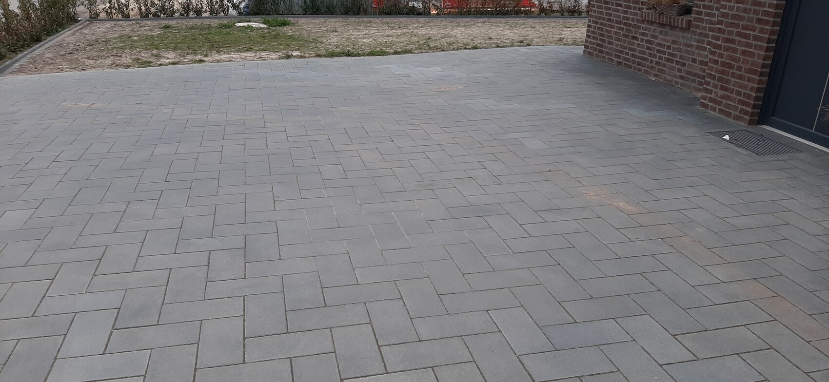 Graue Pflasterfläche aus rechteckigen Steinen im Herringbone-Muster vor einer Backsteinfassade mit Tür und Rost.