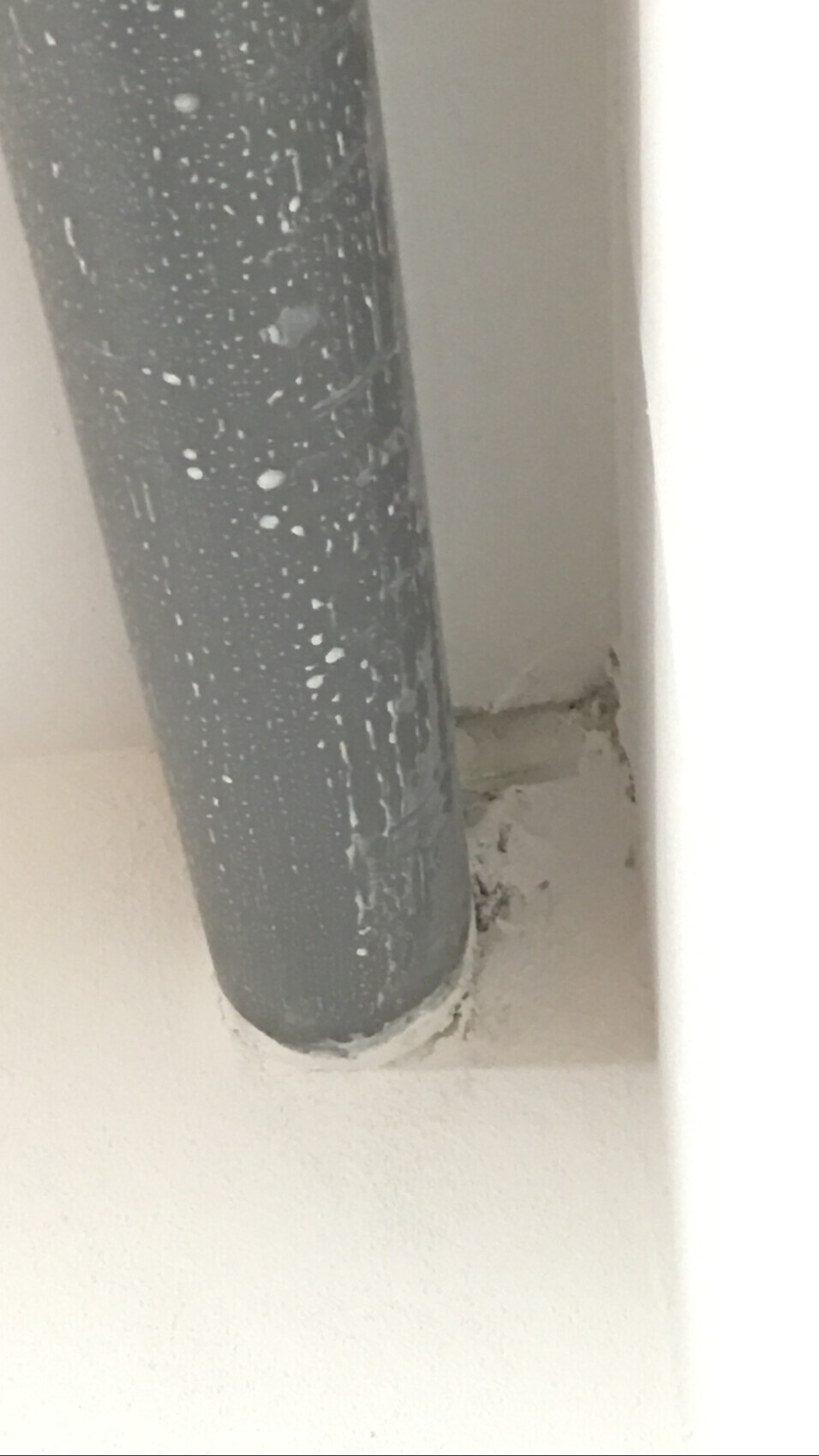 Grau lackiertes Rohr mit weißen Spritzern, an der Wand befestigt, Bodenbereich sichtbar.