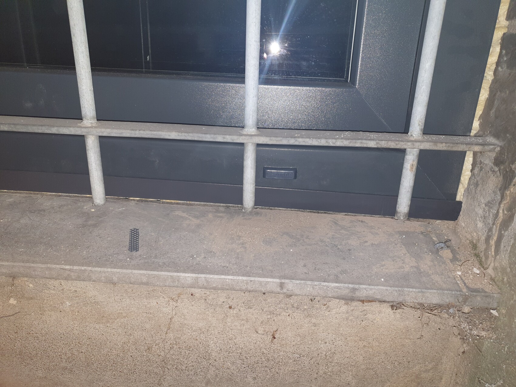 Metallgitter vor dunklem Fensterrahmen; Betonboden und Steinwand im Hintergrund.