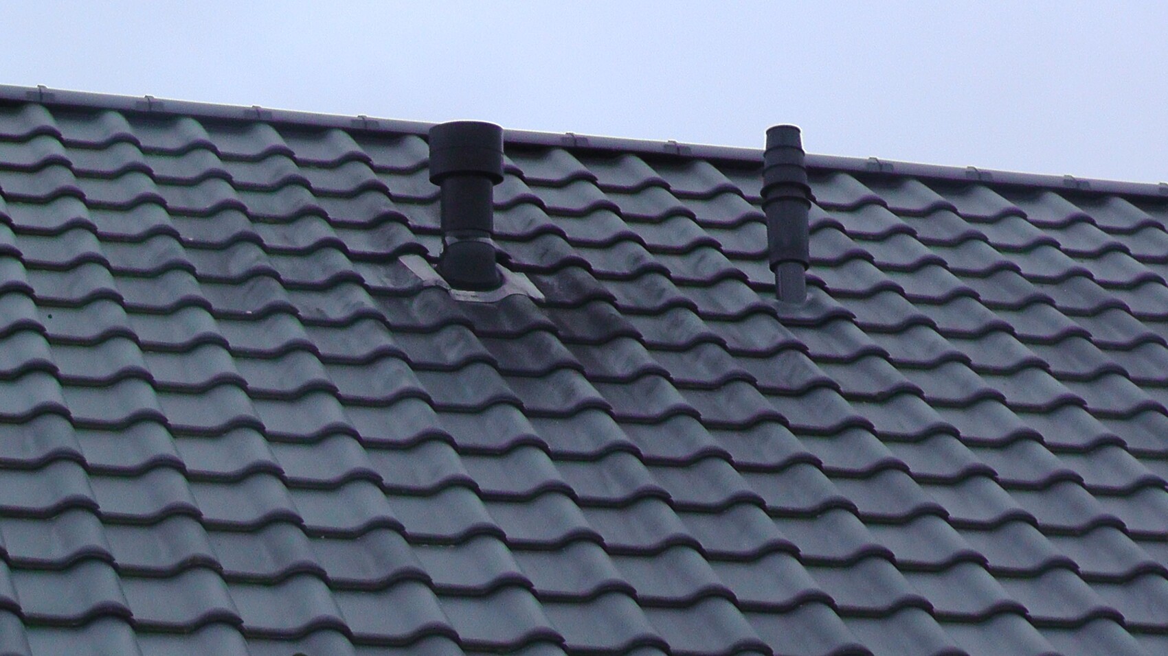 Dach mit grauen Wellenziegeln und zwei schwarzen Kaminrohren gegen blauen Himmel.