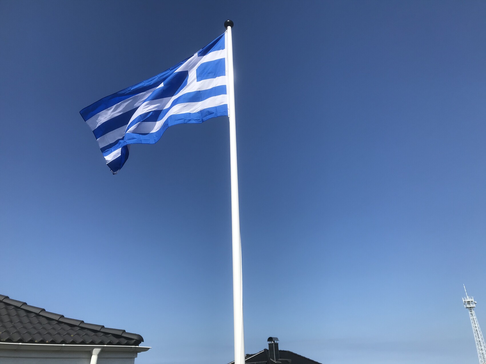Griechische Flagge weht am Mast vor klarem blauem Himmel, Dächer im Vordergrund.