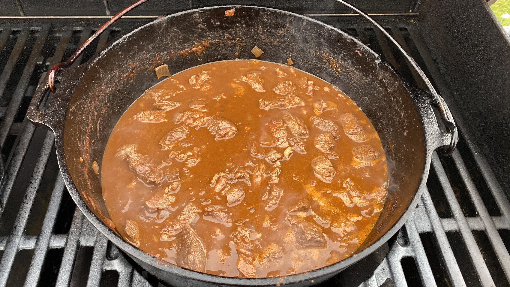 Gusseiserner Topf mit braunem Fleisch-Eintopf auf Grillrosten; Fleischstücke in roter Sauce.
