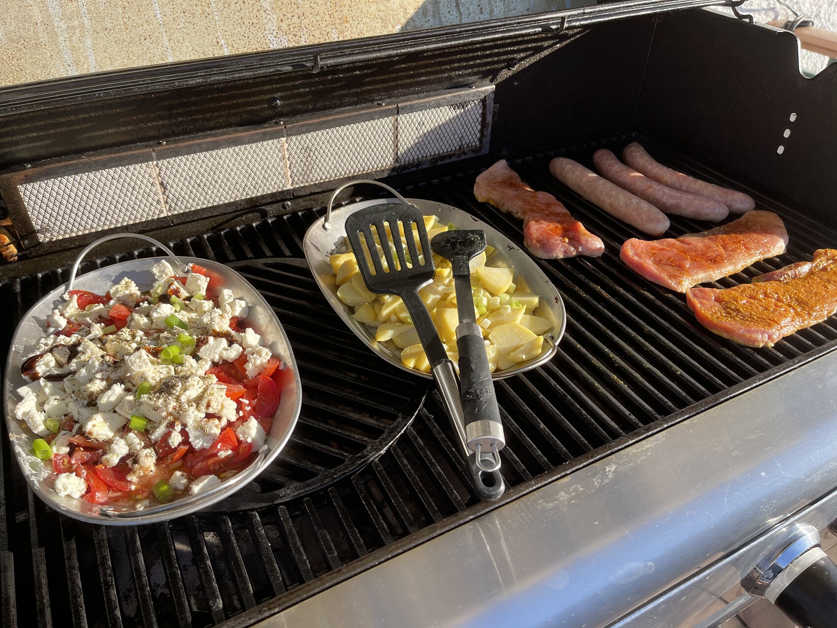 Grillfläche mit rohen Würstchen und Fleisch, daneben Kartoffelstücke und Tomaten-Feta-Salat.