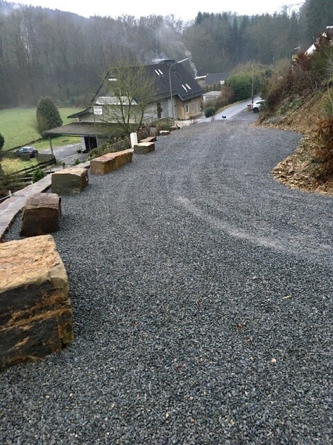 Kiesweg mit Steinblöcken links, Haus mit Kamin am Hang, Wald hinter dem Gebäude.