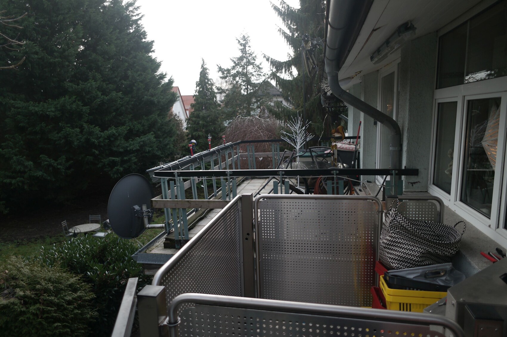 Balkon mit perforiertem Metallgeländer, Satellitenschüssel, Kisten und Gartenzubehör vor Häusern.