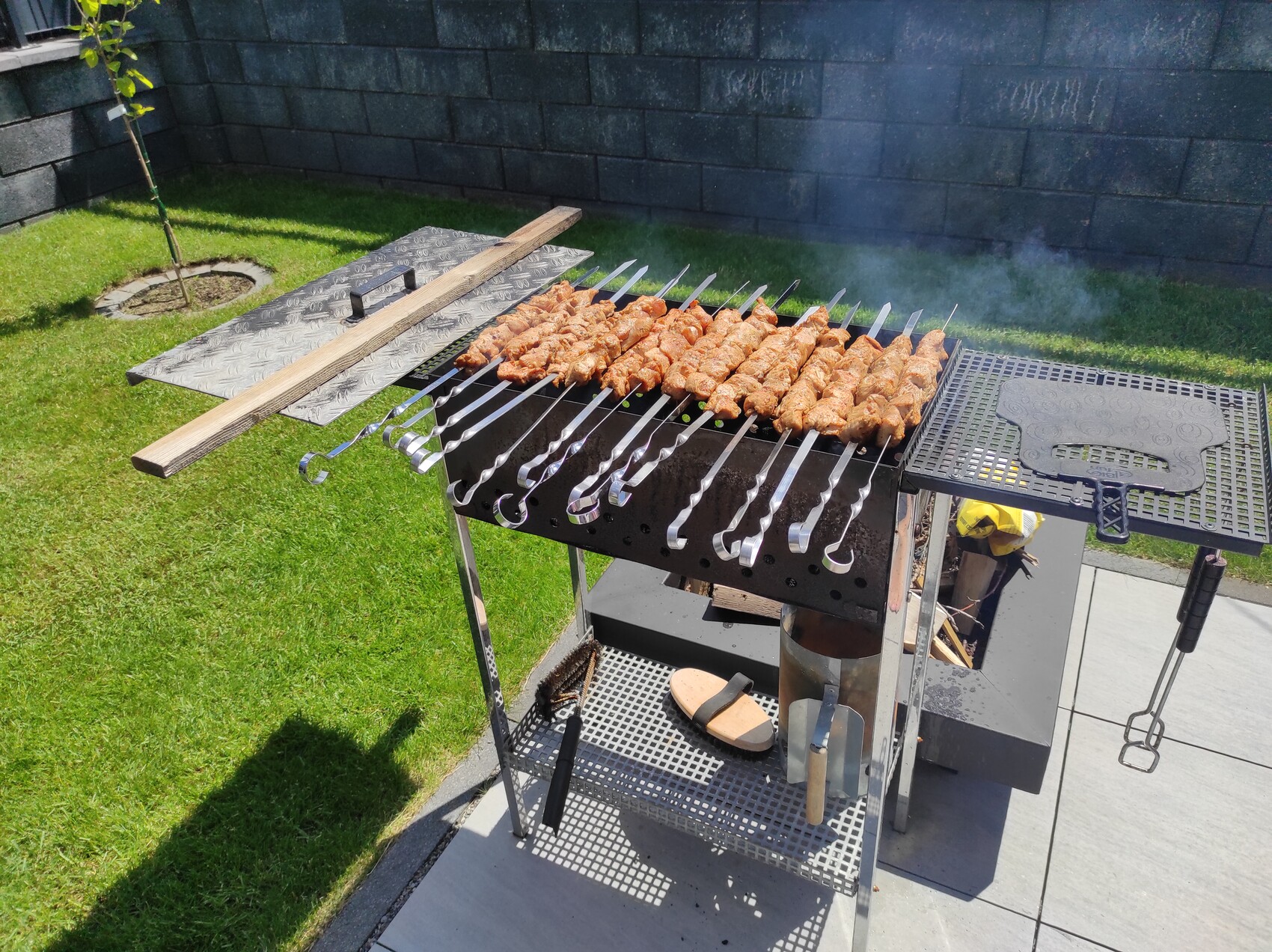 Grill mit Spießen aus Fleisch über Holzkohle im Garten, Rauch steigt.