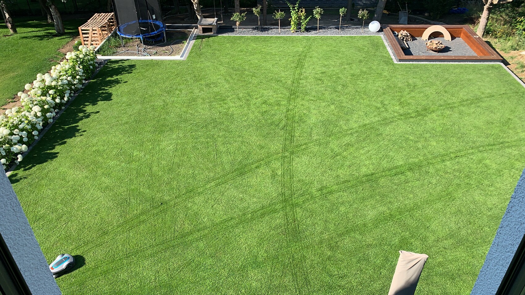 Großer Garten mit grünem Rasen, links Trampolin und Hortensien, rechts Feuerstelle im Holzrahmen.