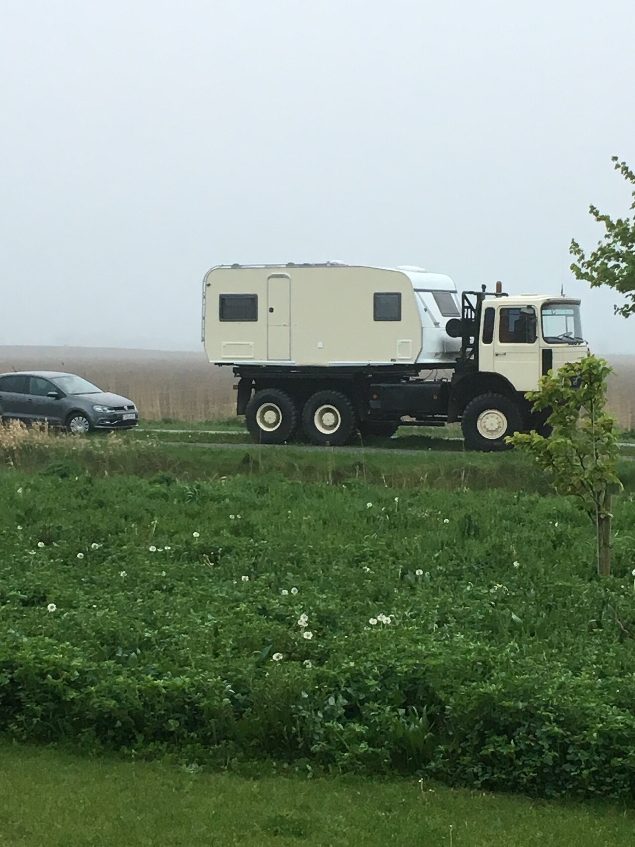 Großes Geländefahrzeug mit beigeem Wohnwagen-Anhänger auf grünem Feld, neben einem Pkw.