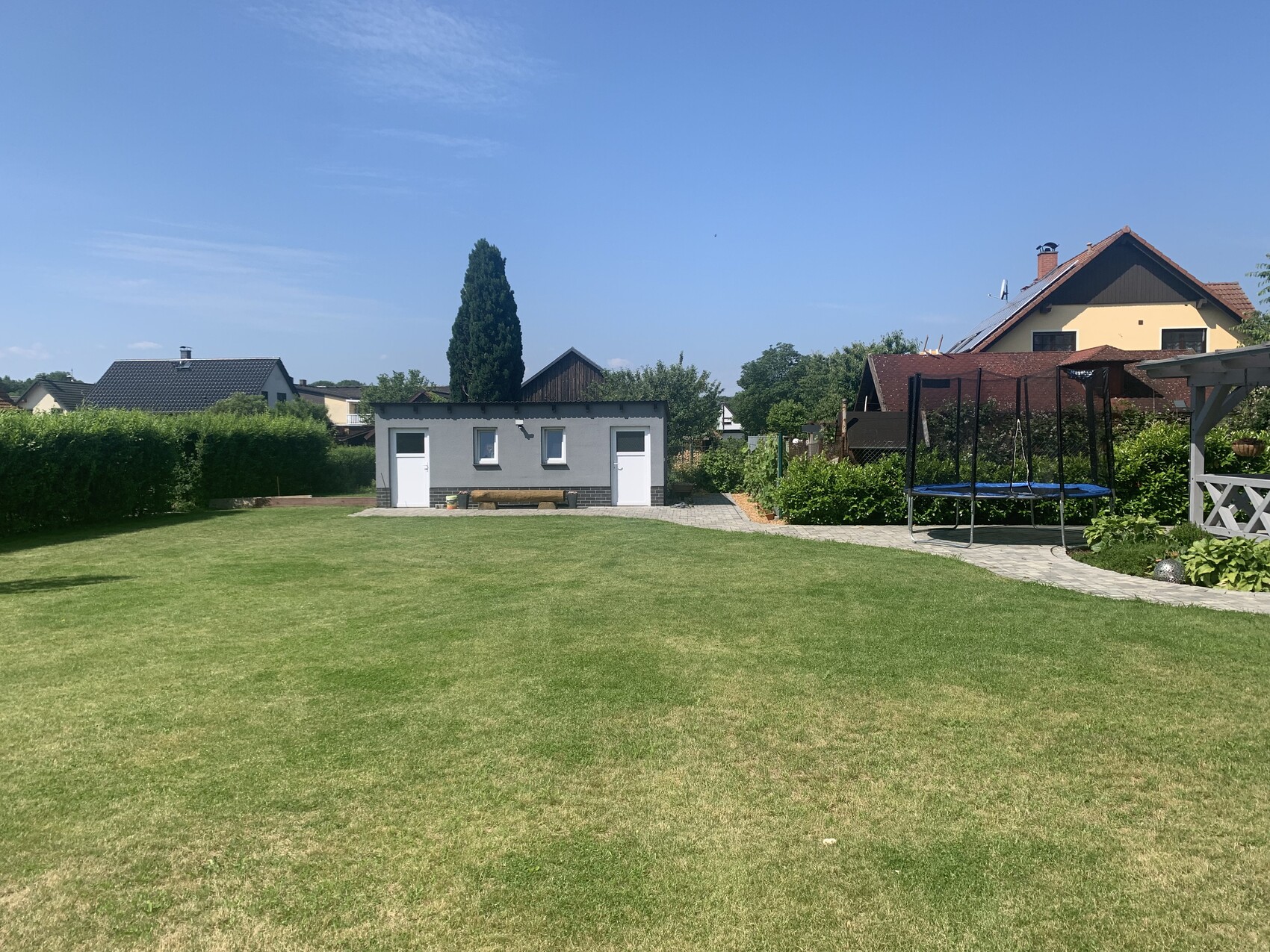 Garten mit grünem Rasen, kleinem grauen Bau, Trampolin rechts, Häuser im Hintergrund.