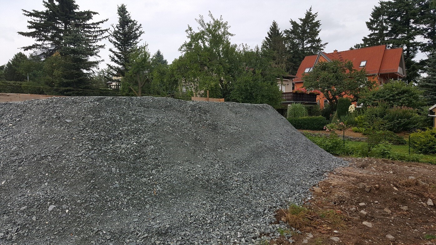 Großer Kieshaufen auf einer Baufläche neben einem Wohnhaus mit Garten