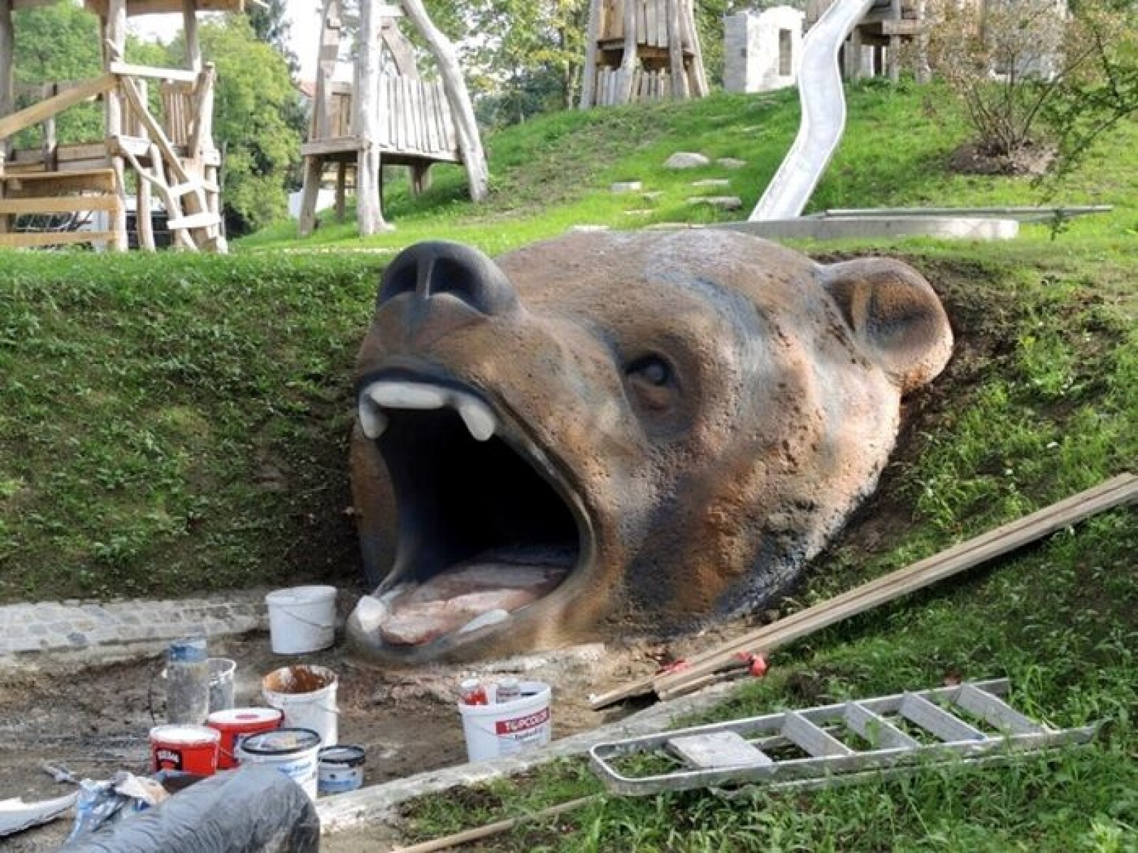Großer Bärenkopf-Skulptur, halb im Erdreich, Farbtöpfe und Leiter vor Spielplatz im Hintergrund.