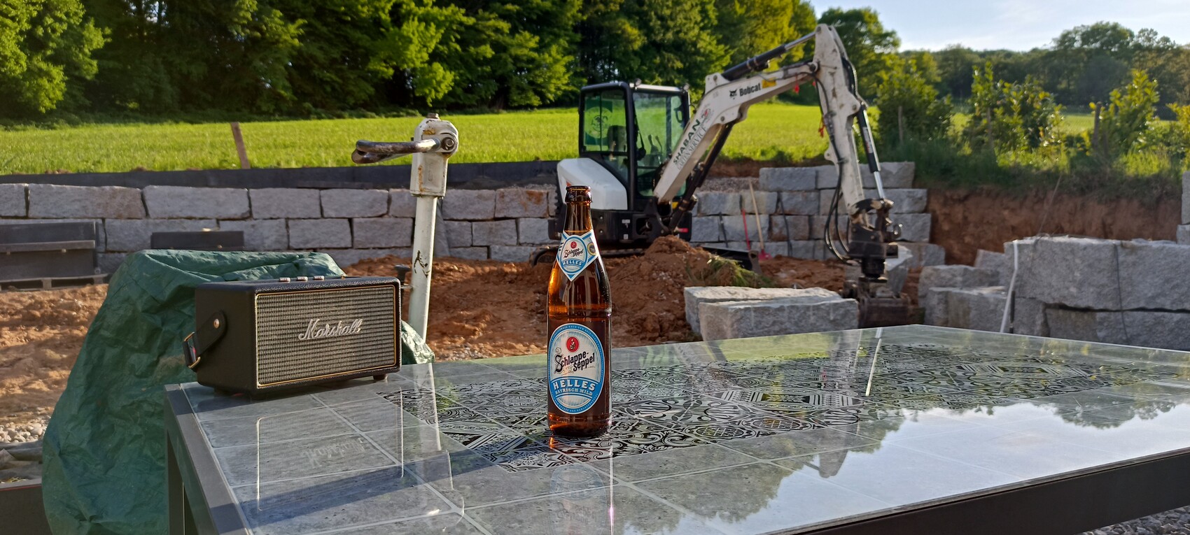 Bierflasche steht auf glänzender Metall-Tischplatte, daneben Bauplatz mit Bagger und Steinblöcken.