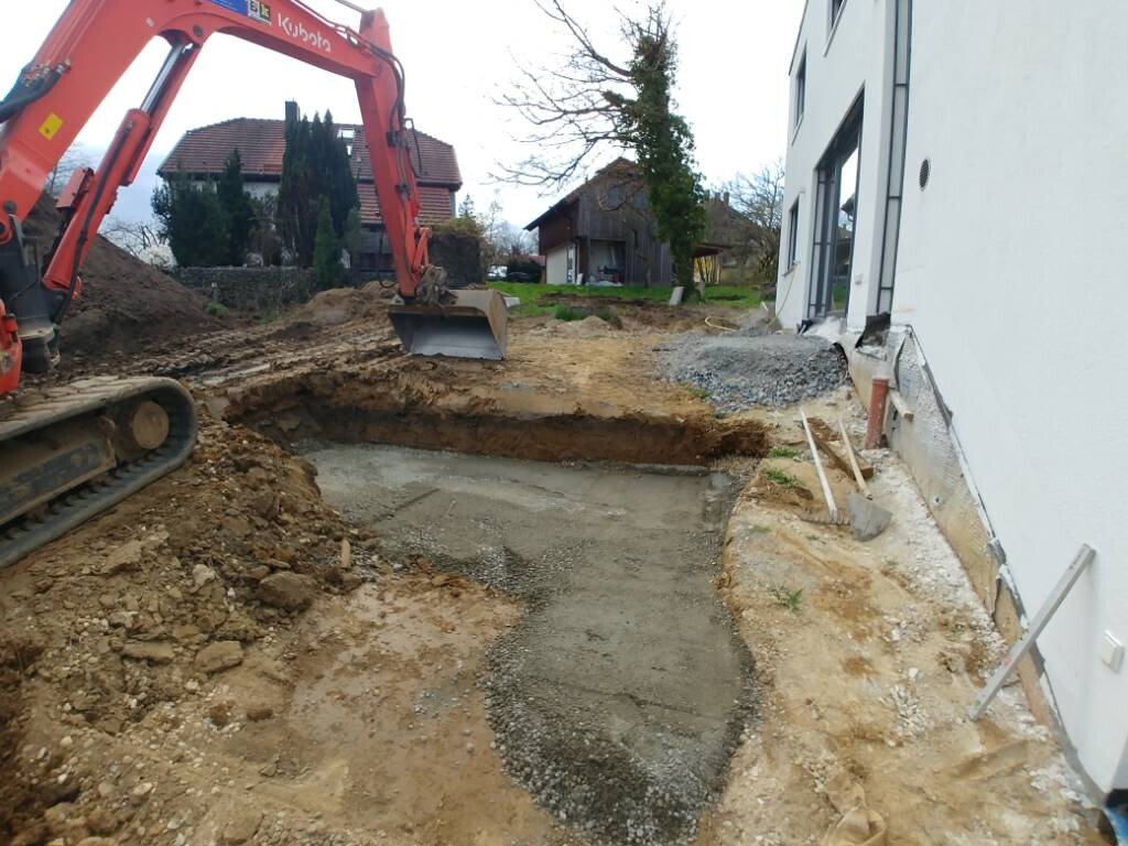 Roter Bagger gräbt eine Baugrube neben einem weißen Haus; Erdaushub und Kies liegen bereit.