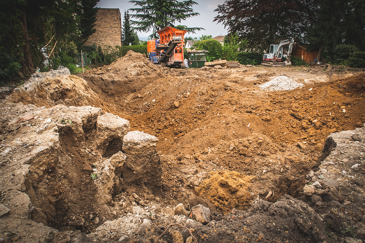 Große Aushubgrube auf Baustelle mit Erdreichhaufen und Baggern im Hintergrund.