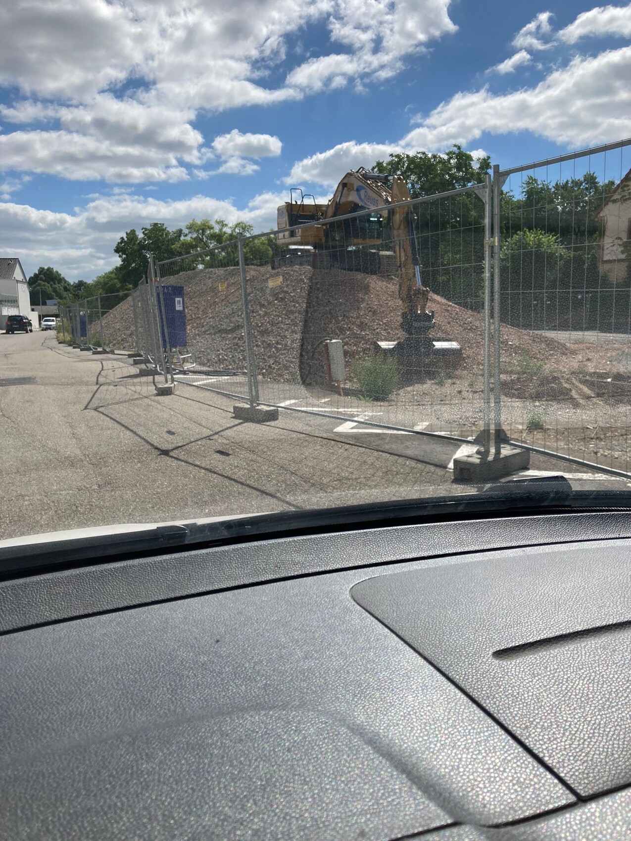 Baustelle am Straßenrand: Bagger auf Erdhaufen hinter Bauzaun, Sicht aus dem Auto.