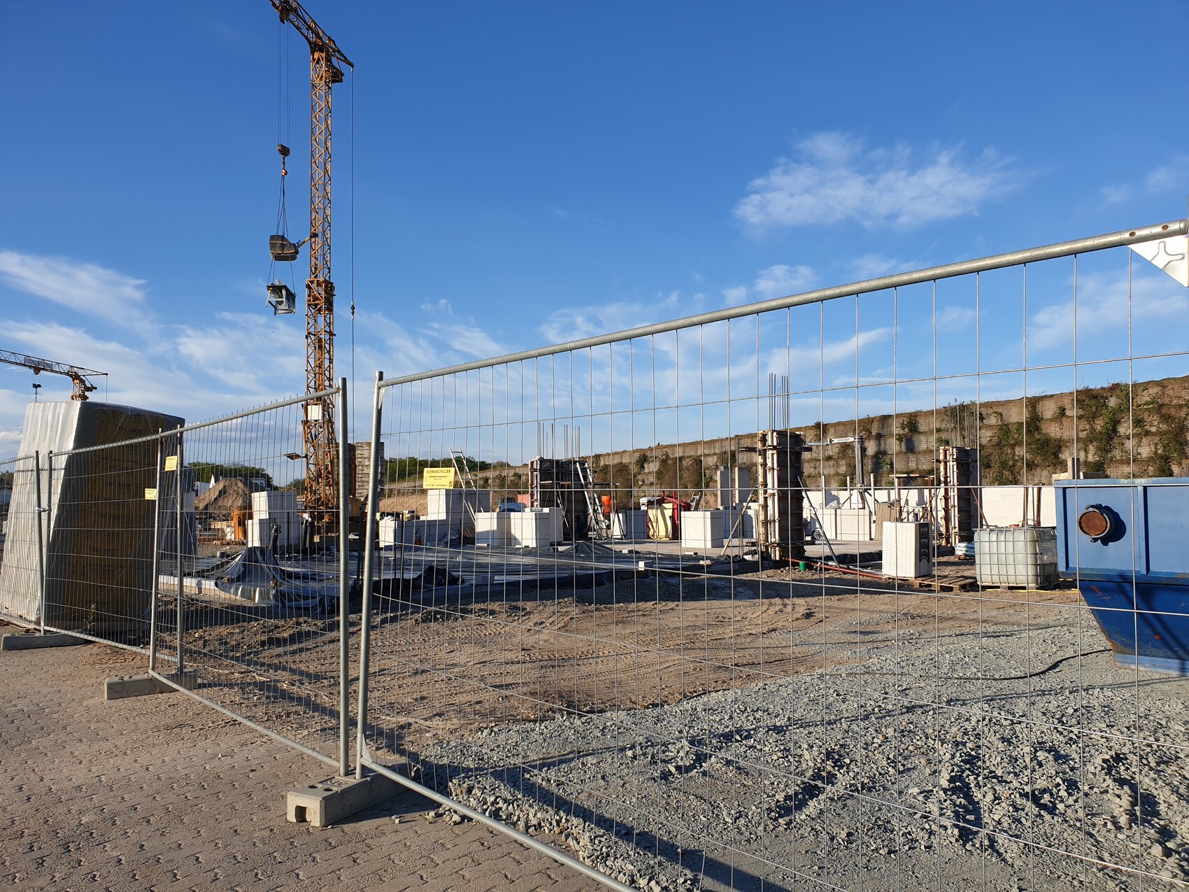 Baustelle mit Kran hinter Metallzaun, Baumaterialien und Baugeräten, blauer Himmel.