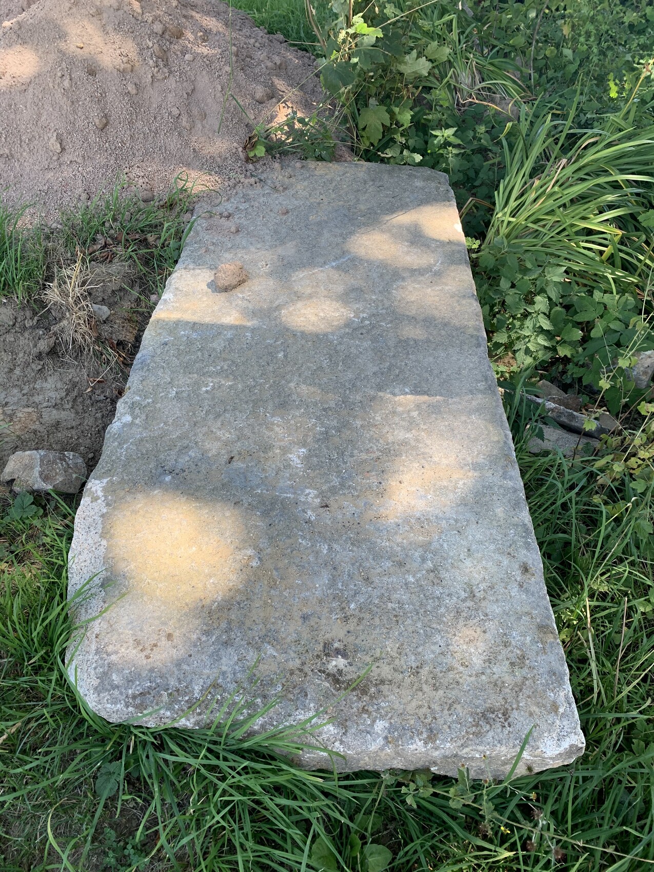 Grobe Betonplatte liegt im Gras neben einem Erdhügel, von Pflanzen umgeben.