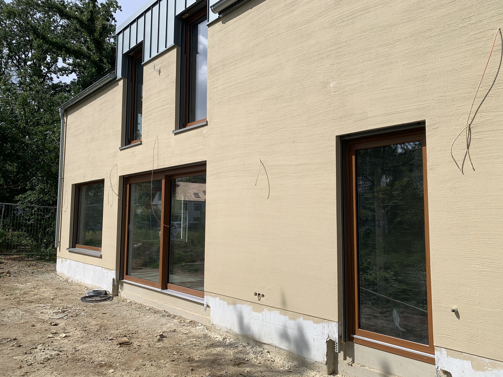 Außenansicht eines Neubauhauses: beigefarbene Fassade, große Fensterfronten, unbefestigter Boden.