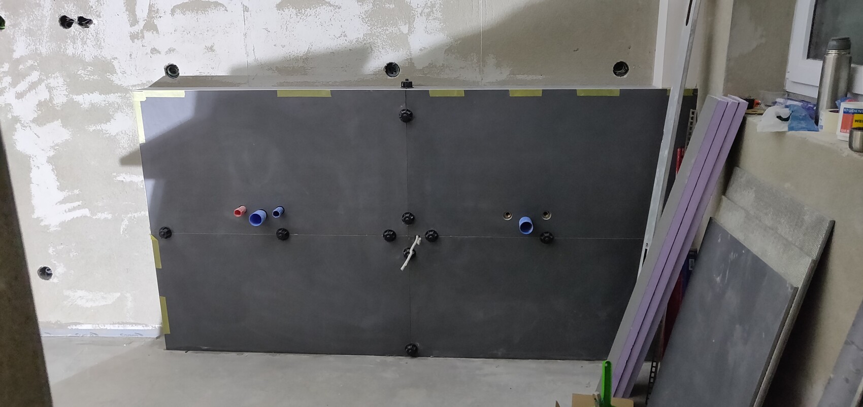 Große dunkle Wandpaneele mit Rohranschlüssen, Schrauben und Klebeband in einer Baustelle.
