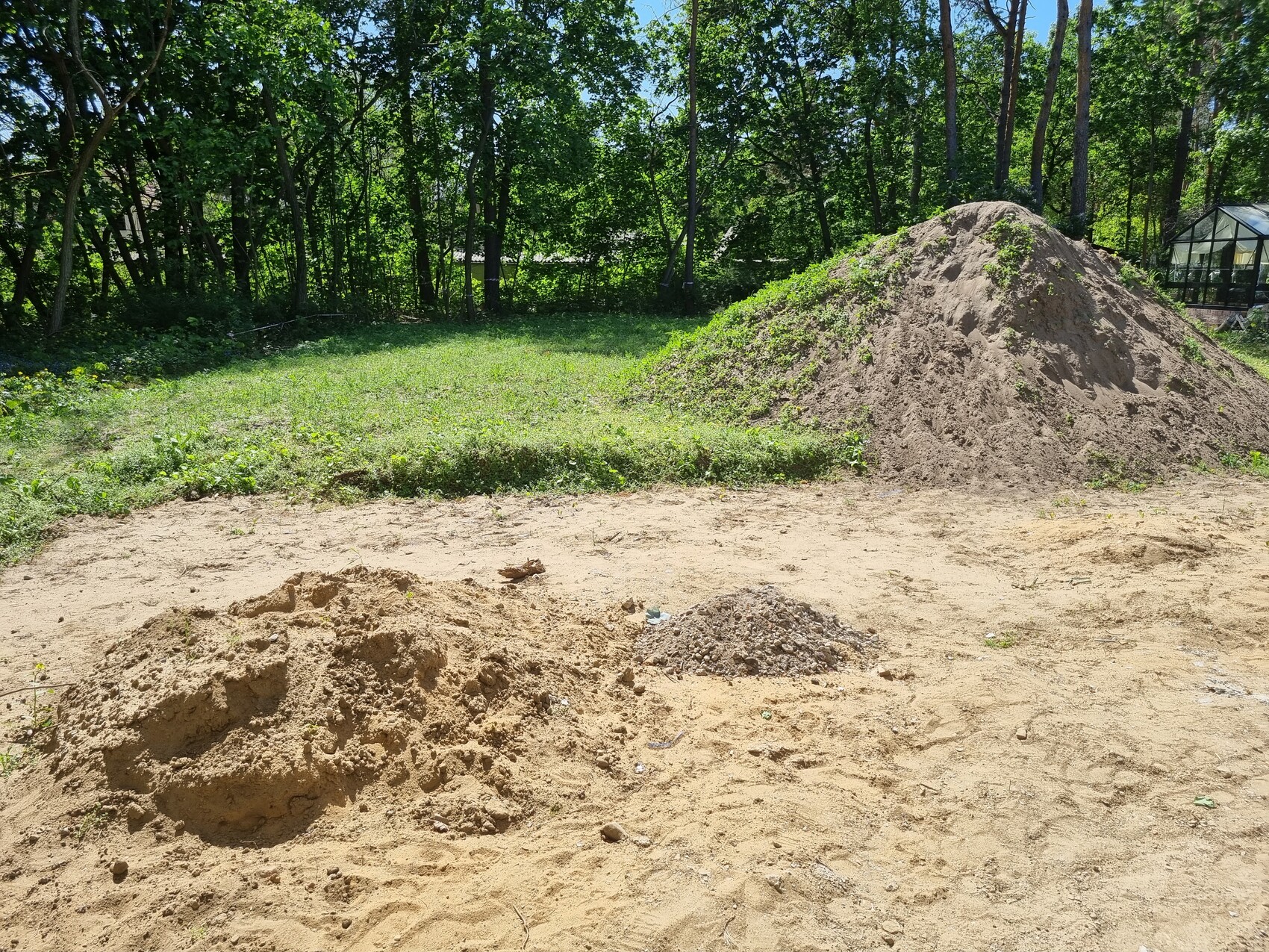 Großer Erd- und Sandhaufen auf freier Fläche, Hintergrund aus Bäumen, rechts Glas-Gewächshaus.