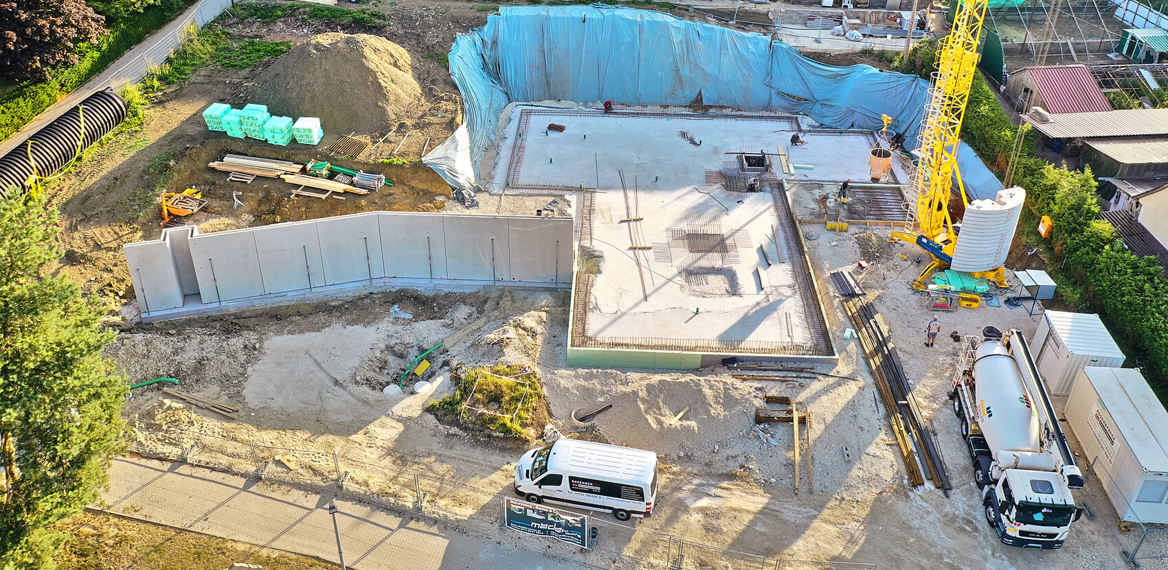 Luftaufnahme einer Baustelle: Fundamentarbeiten, Kran, Betonmischer, Baumaterialien und Blauplane.