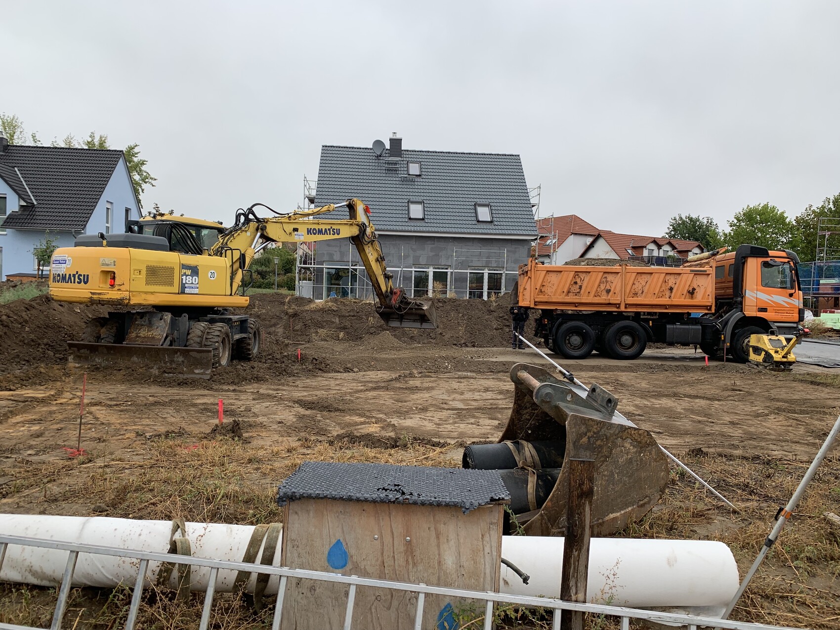 Baustelle mit gelbem Komatsu-Bagger und orangeem Kipper, Häuser im Hintergrund.