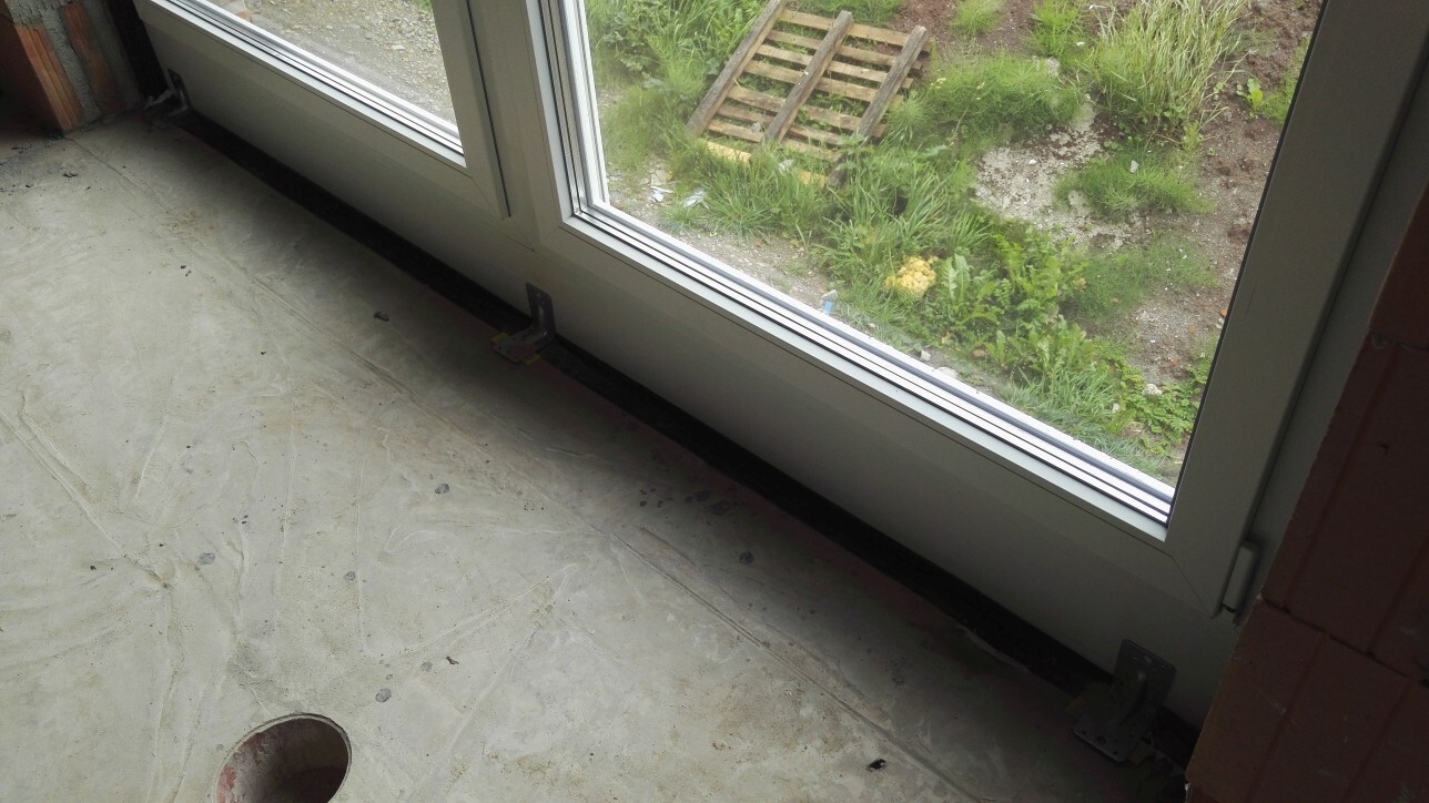 Blick auf eine große Glas-Schiebetür im Rohbau, Betonboden und Gartenbereich außerhalb sichtbar.