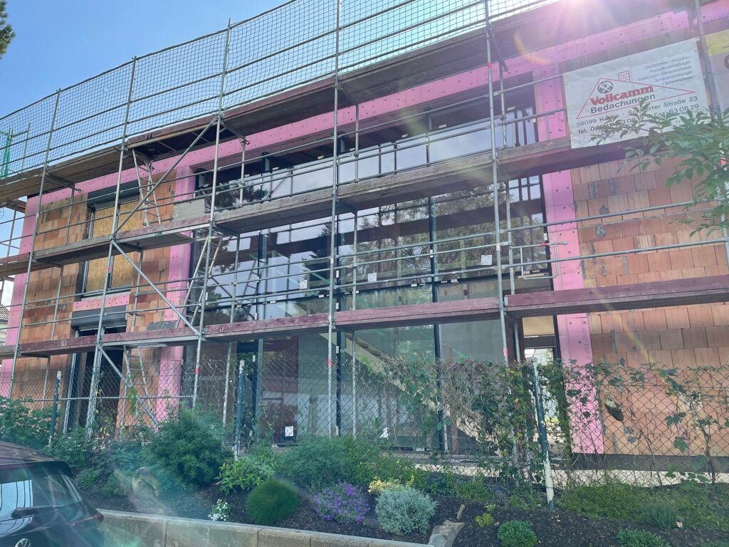Baustelle an modernem Gebäude: Gerüst, große Glasfronten, pinke Isolierung, Garten davor.