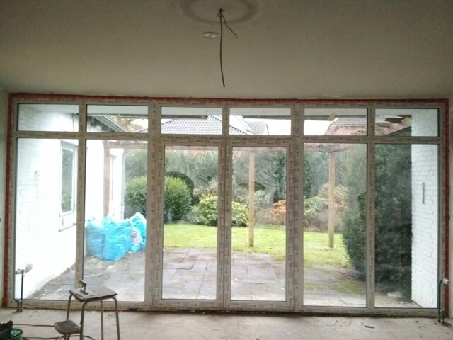 Unfertiger Innenraum mit bodentiefen Glasfenstern zum Garten; Decke offen, Baumaterialien.