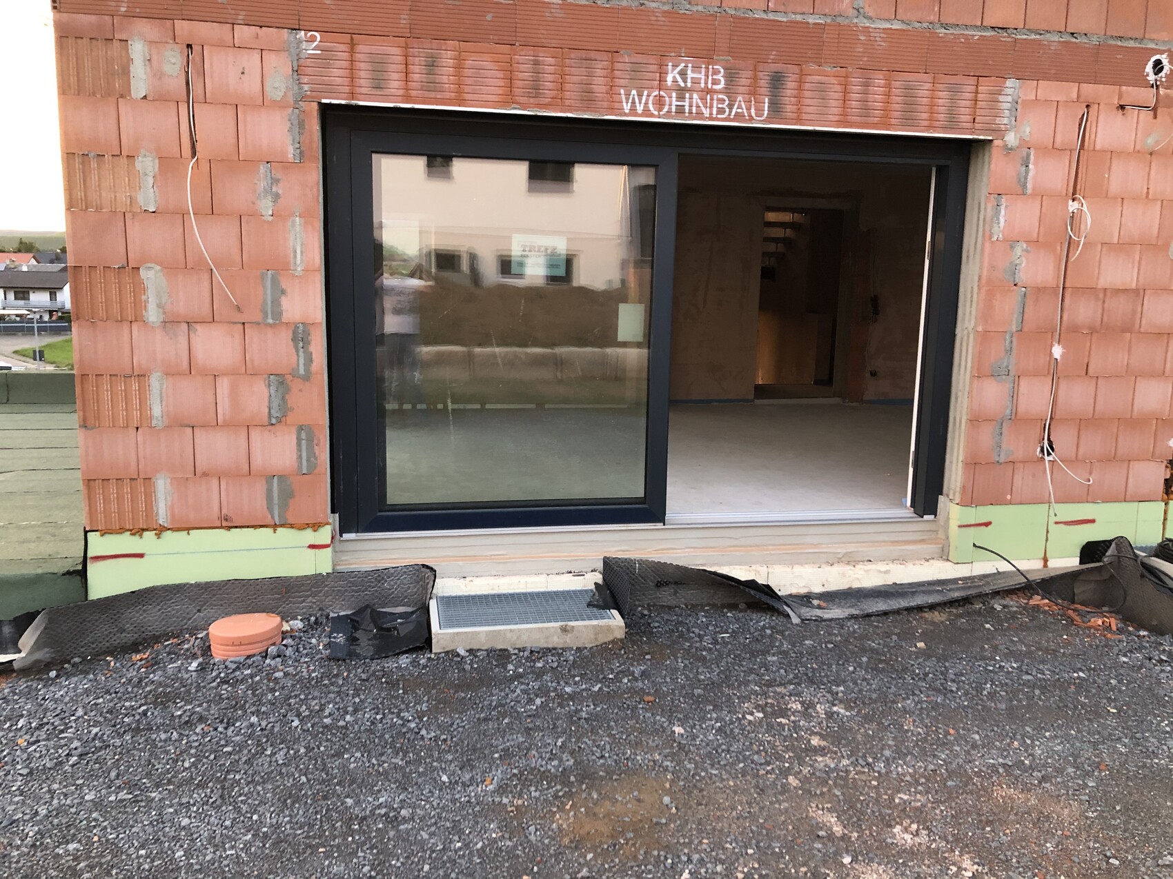 Unfertige Gebäudefassade aus roten Ziegeln mit großer Glas-Schiebetür, Baustelle und Kiesboden.