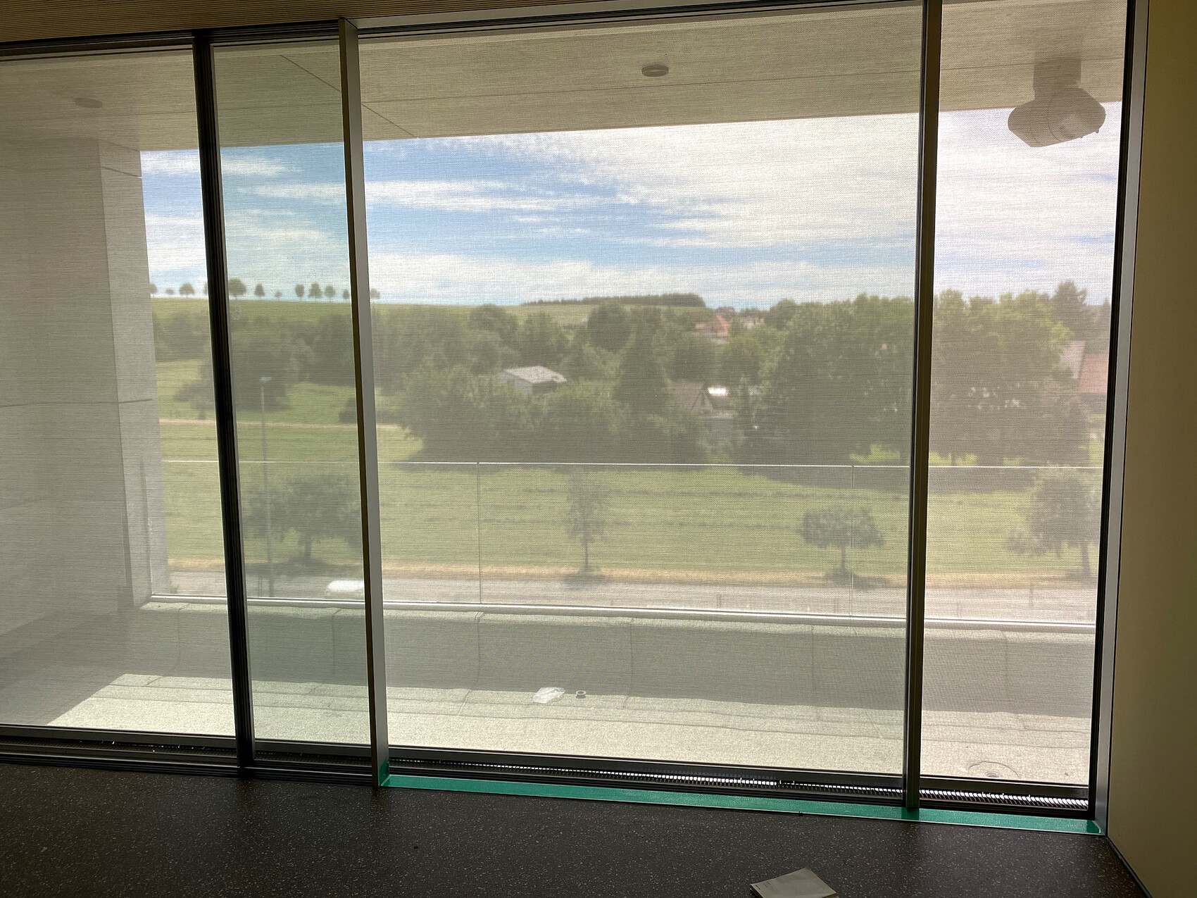 Großes Glasfenster: Blick auf grüne Felder, Bäume und blauen Himmel; Innenraum mit hellen Wänden.