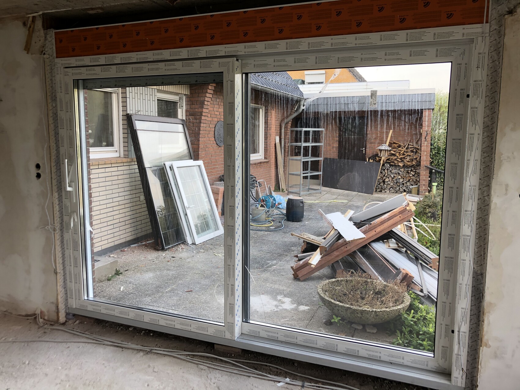 Große Glas-Schiebetür von innen sichtbar; Außen Holzstapel, Metallregale und Bauholz liegen herum.