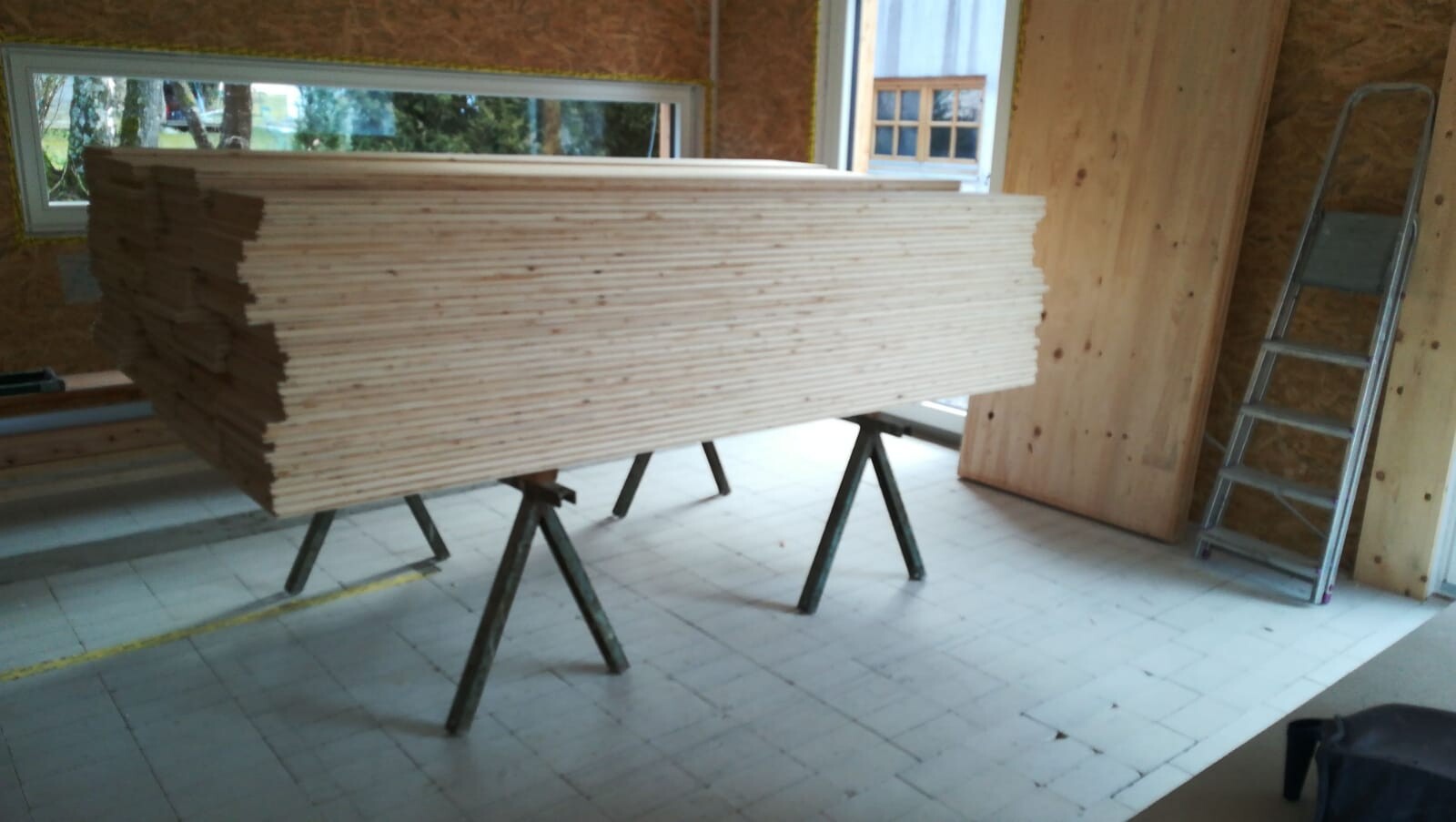 Stapel heller Holzlatten auf zwei Sägeböcken in einer Werkstatt, daneben eine Leiter.