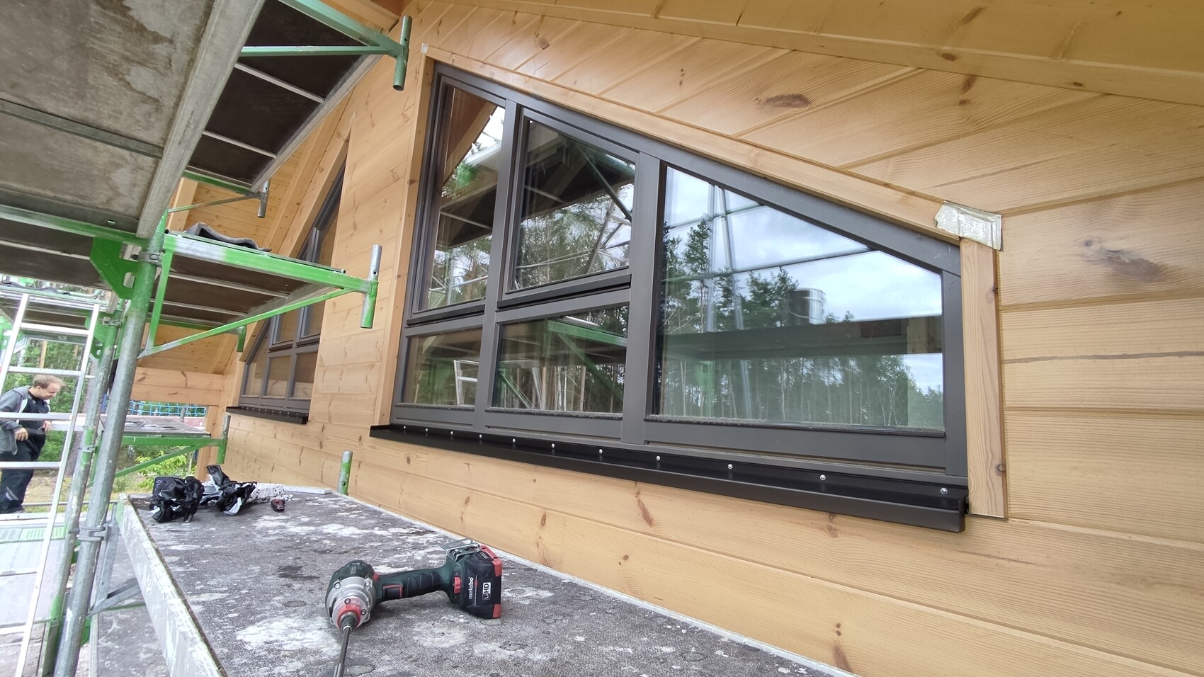 Holzhaus-Baustelle mit großen grauen Fensterrahmen, Metallgerüst und Werkzeugplattform.