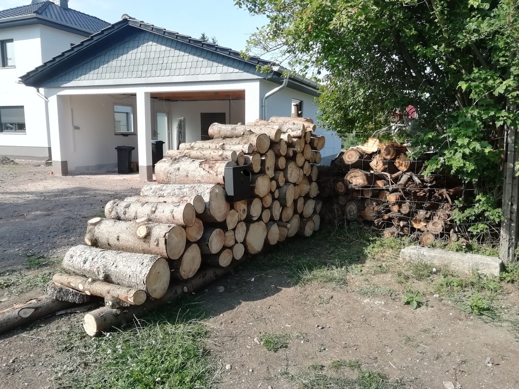Großer Holzstapel aus Baumstämmen vor weißem Einfamilienhaus mit Vorgarten.