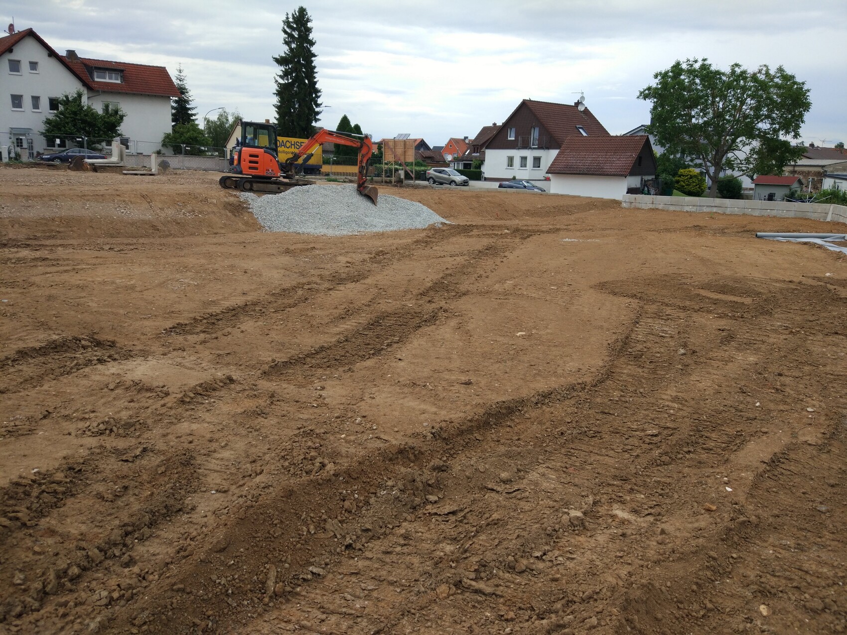 Baugrundstück mit Bagger, Kieshaufen und Wohnhäusern im Hintergrund.