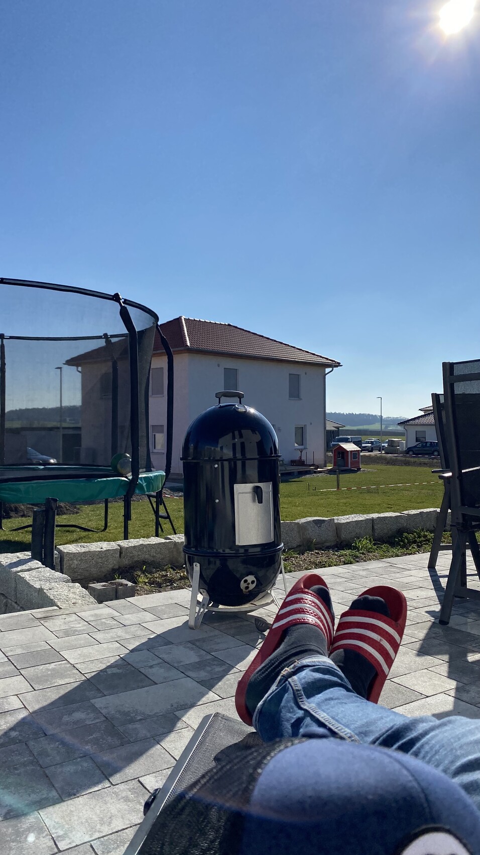 Garten-Terrasse: Person entspannt auf Liege, roten Schuhen; Grill, Trampolin, Haus im Hintergrund.