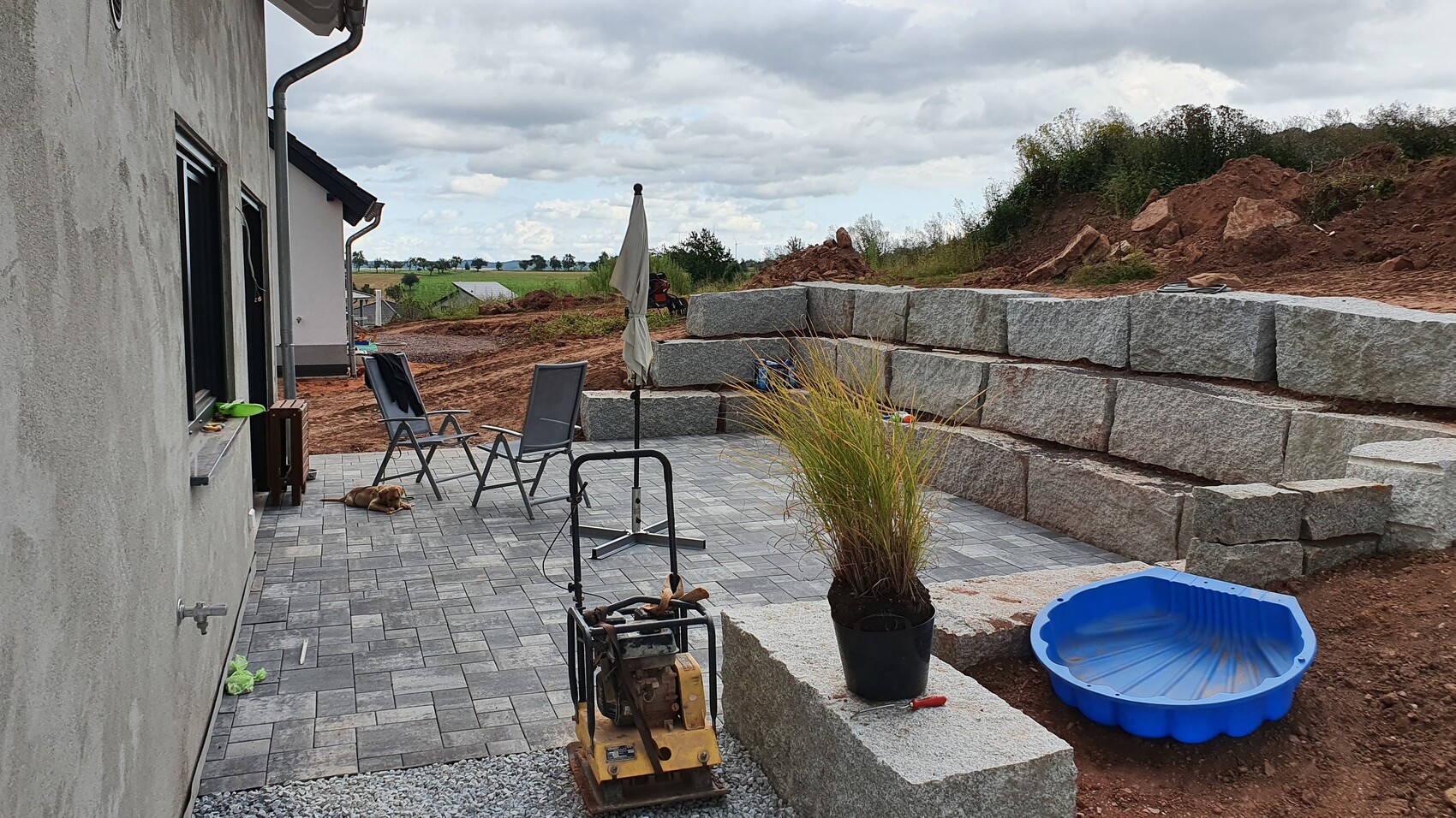 Garten-Patio mit Lounge-Stühlen, geschlossenem Sonnenschirm, Hund liegt, Steinmauer.