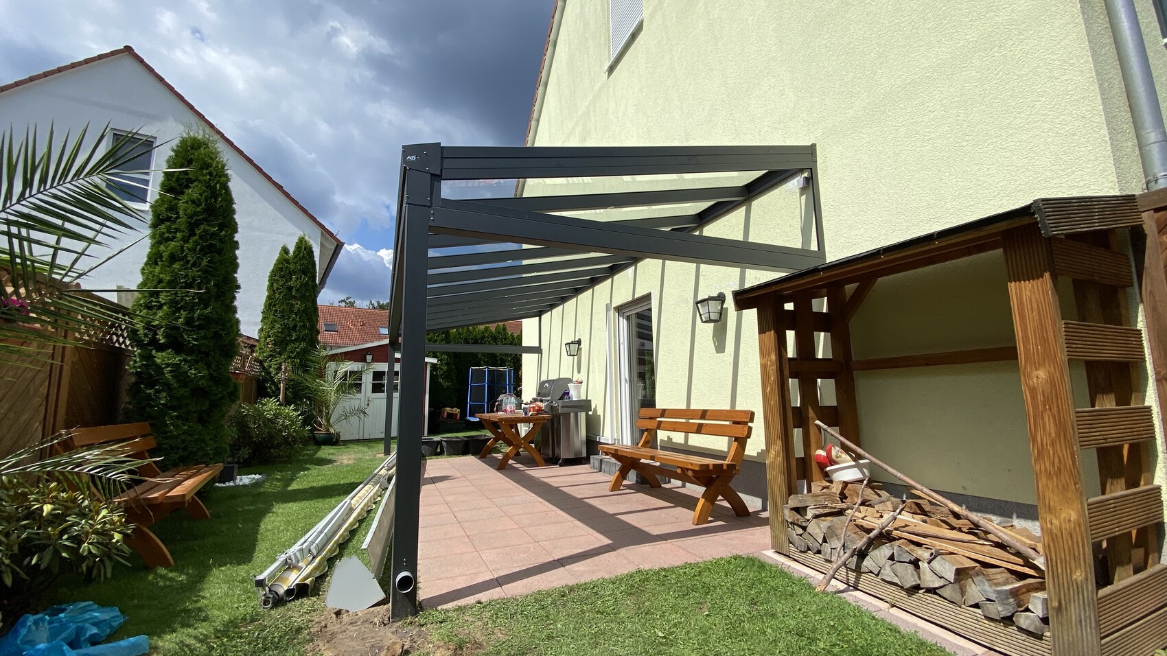 Gartenterrasse mit grauer Metallpergola, Holzbänken, Grill und Brennholz.