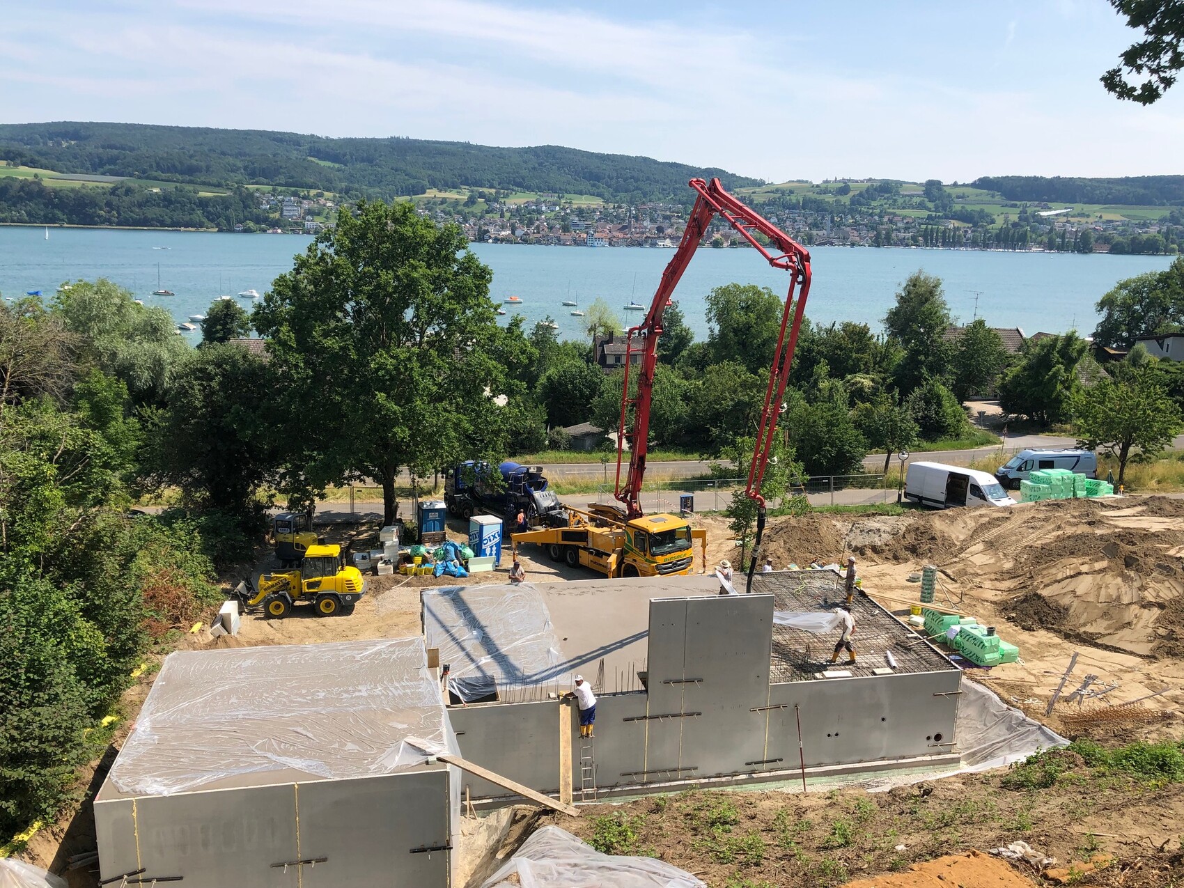 Baustelle am Seesufer: Fundamentmauern, roter Betonpumpenarm, Baugeräte und Arbeiter.
