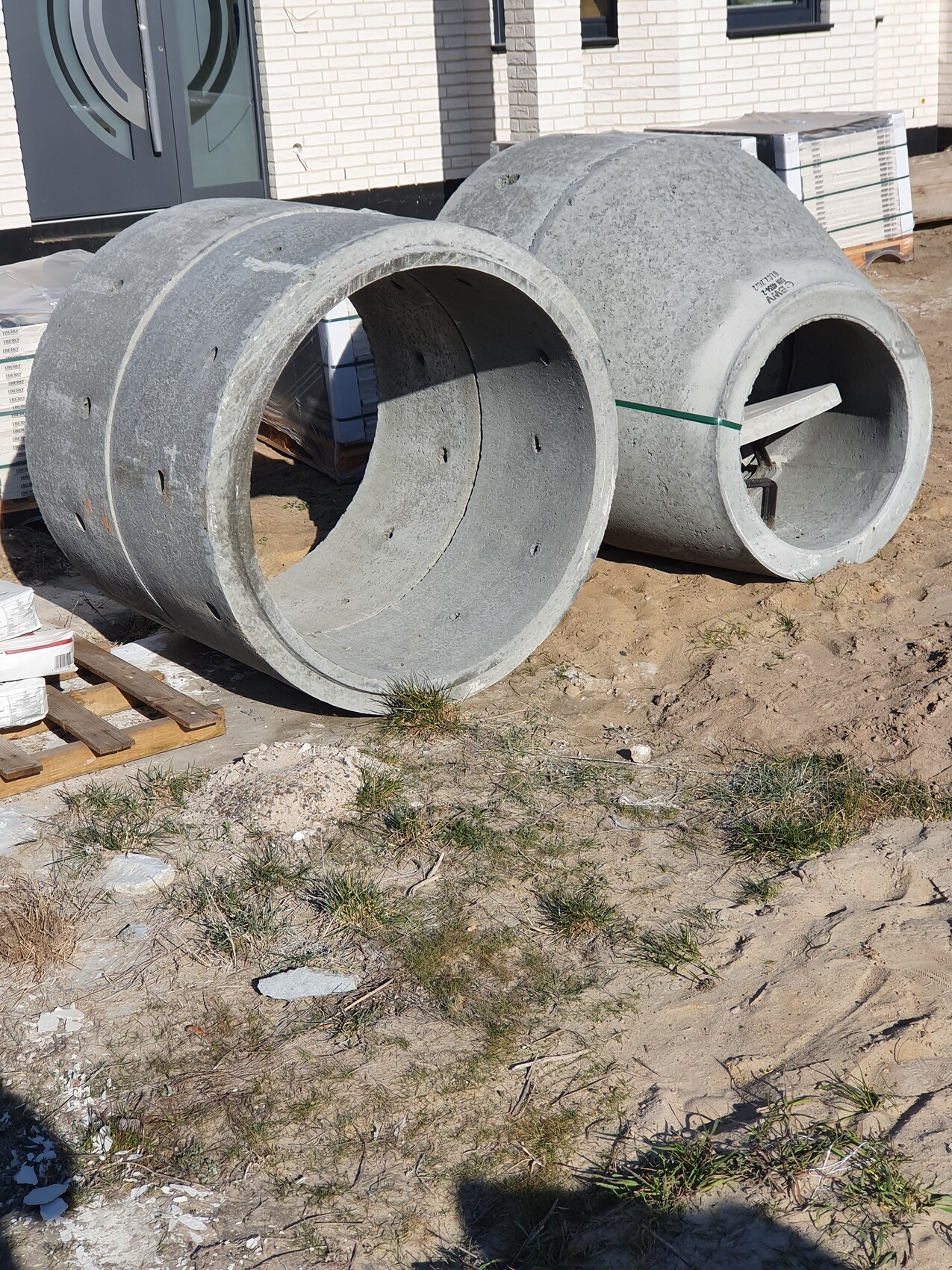 Zwei große graue Betonrohre liegen auf einer Baustelle vor einem Neubau, mit Baumaterialien daneben.