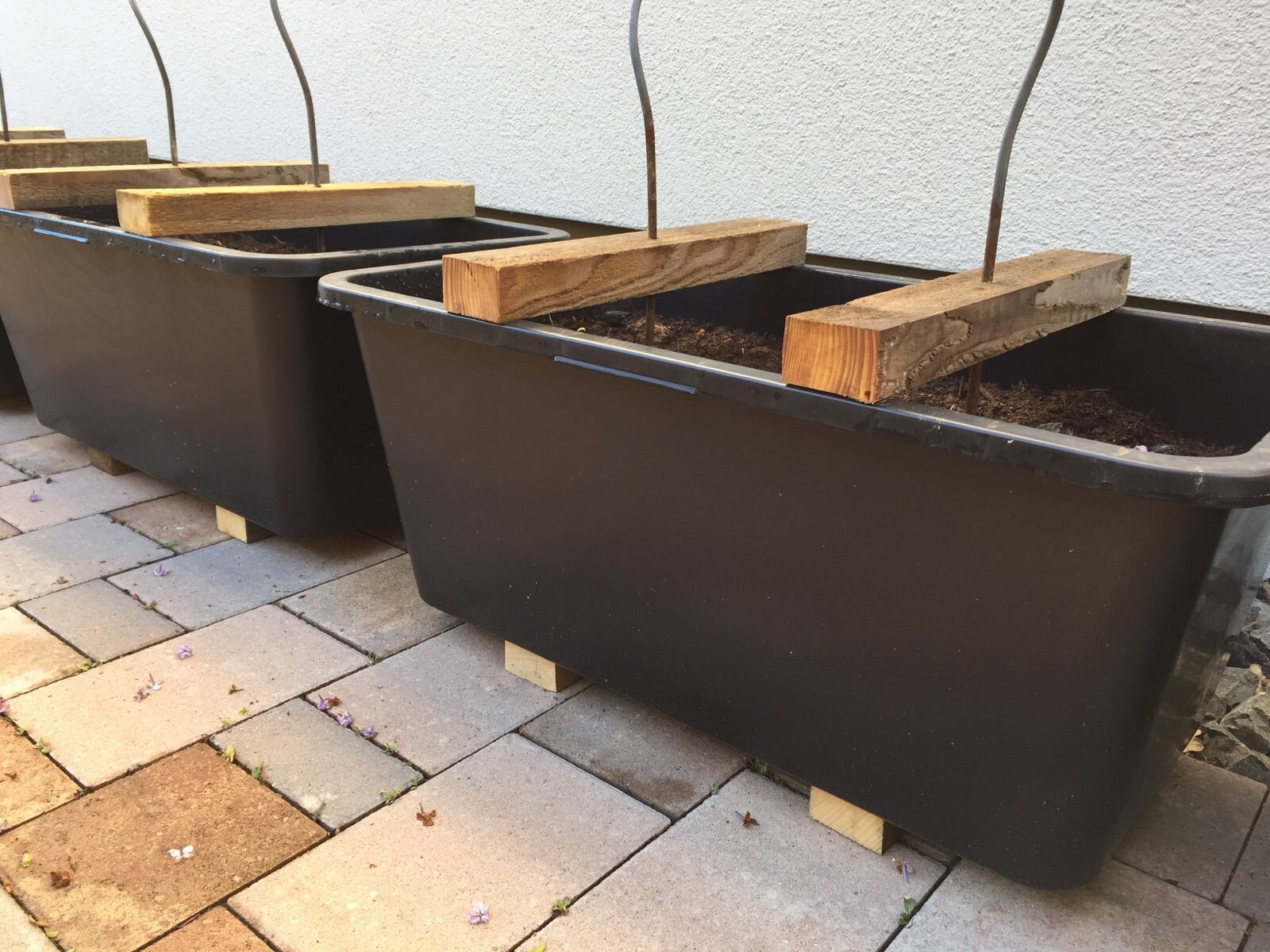 Drei schwarze rechteckige Kunststoff-Pflanzkübel mit Holzbrettern darauf, auf Pflaster neben Wand.