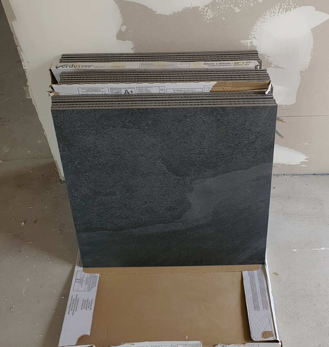 Dunkler Fliesenstapel in offener Kartonschachtel, Wand im Hintergrund, Verpackung sichtbar.