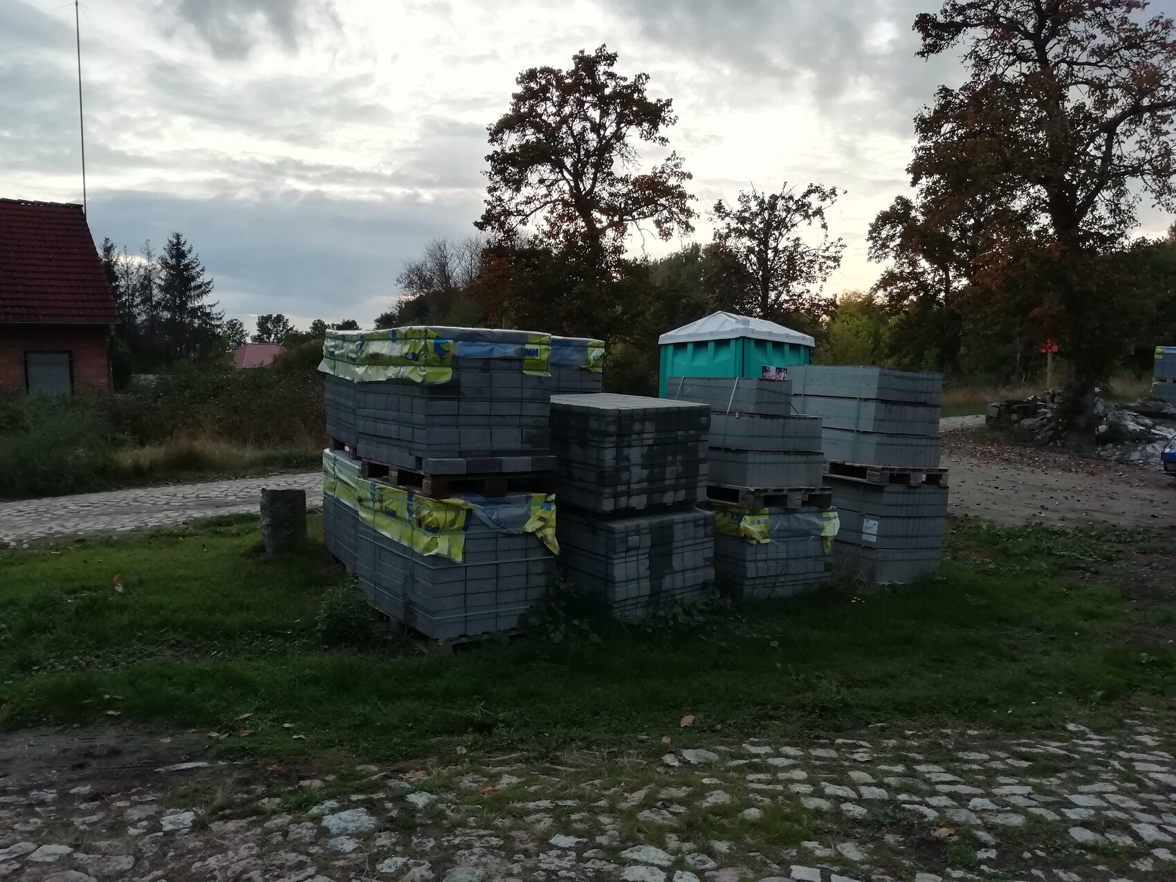 Stapel Betonplatten auf Paletten neben einer mobilen Toilette, Baumreihen im Hintergrund.