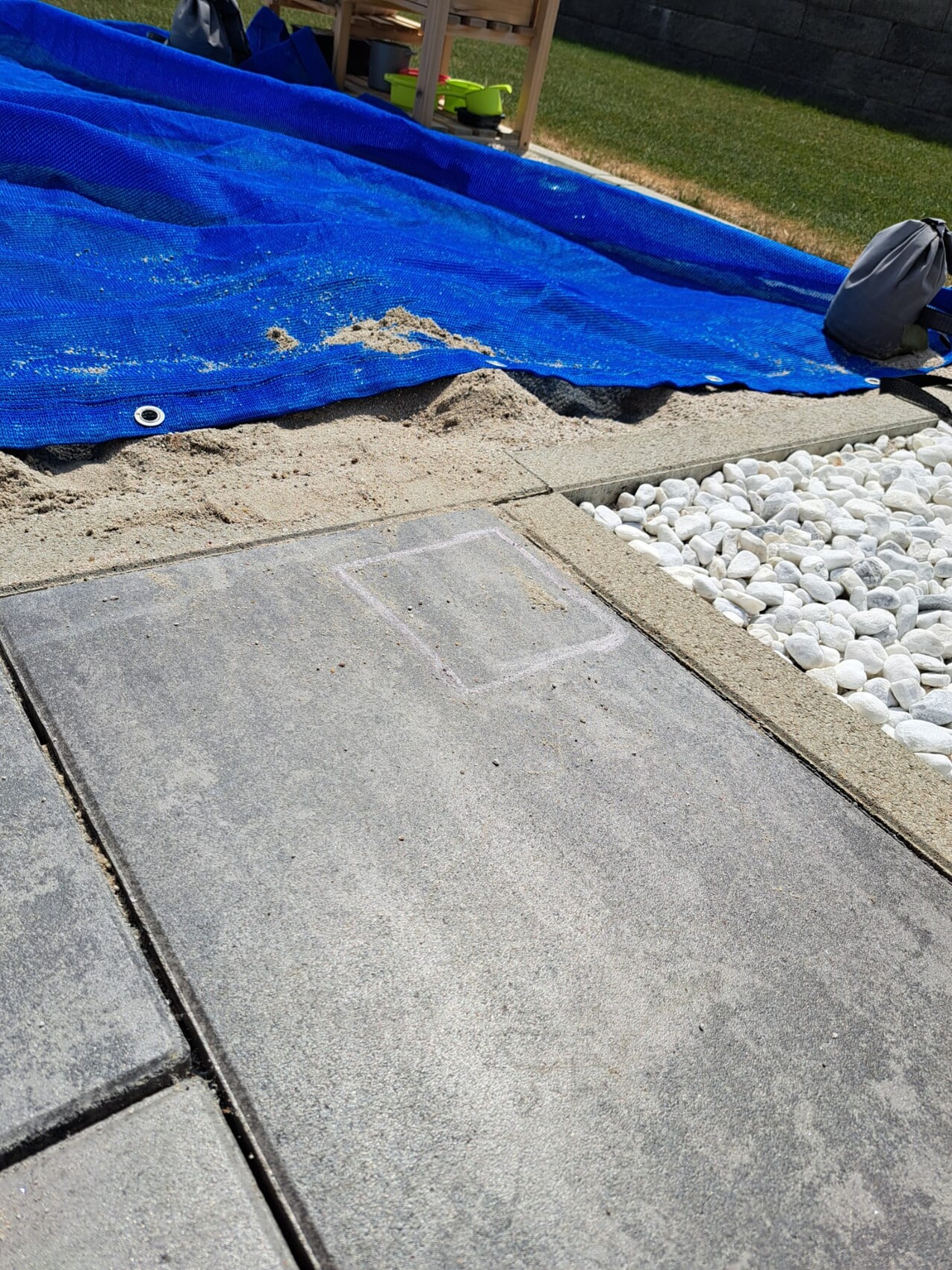 Gartenbereich mit großen Betonplatten, unter blauer Plane liegt Sand; weiße Kiesel am Rand.