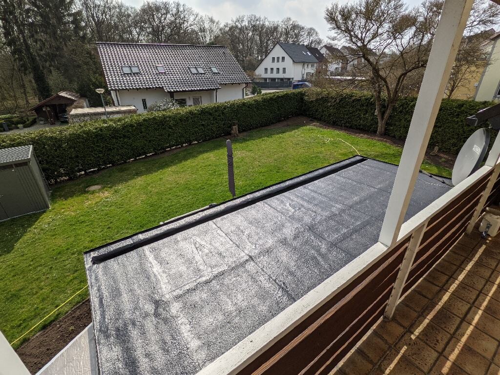Blick vom Balkon auf einen grünen Garten, von einer dichten Hecke begrenzt, mit Häusern dahinter.