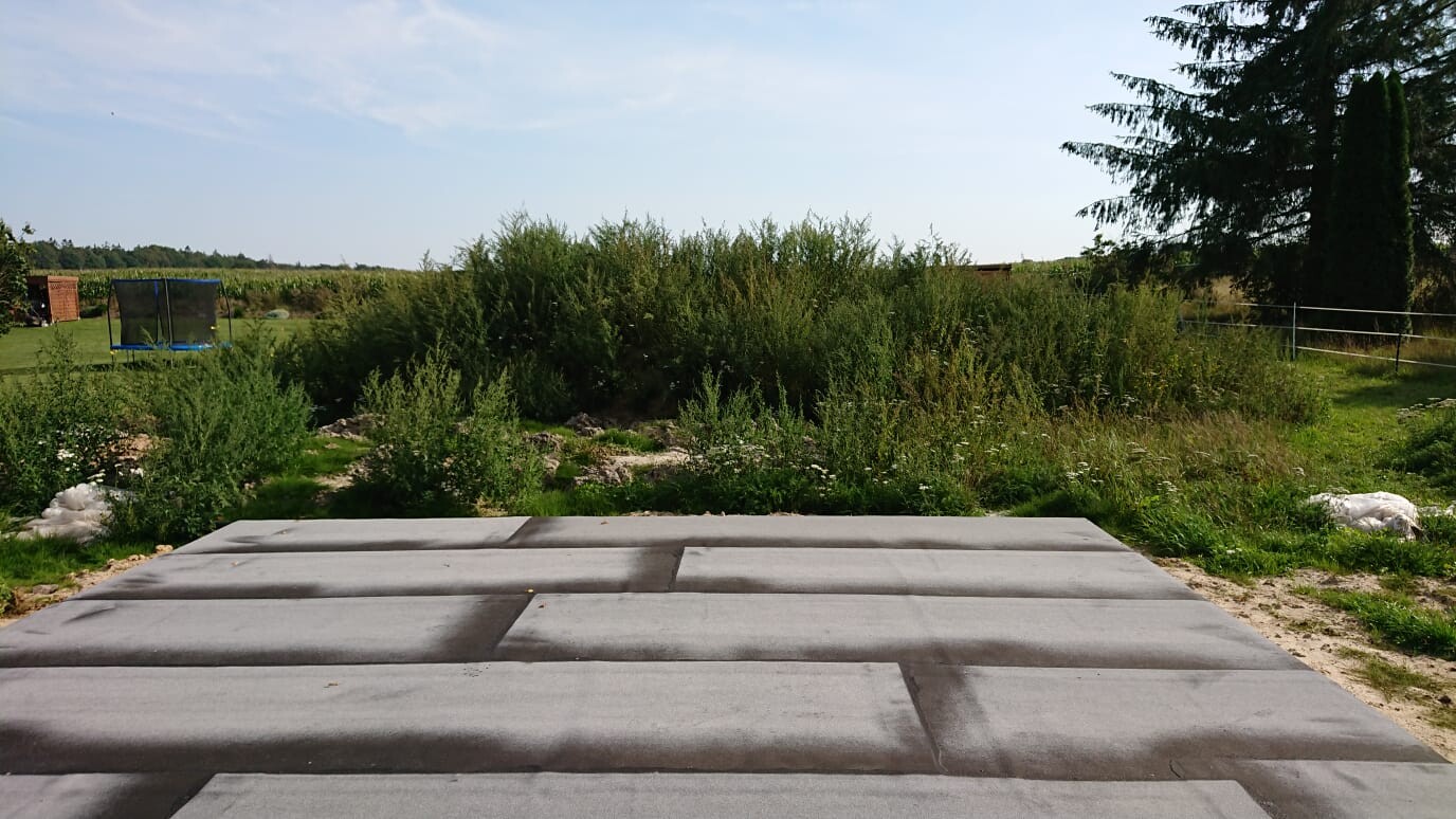 Große graue Betonplatten im Vordergrund, dahinter Buschwerk, Trampolin links und offenes Feld.